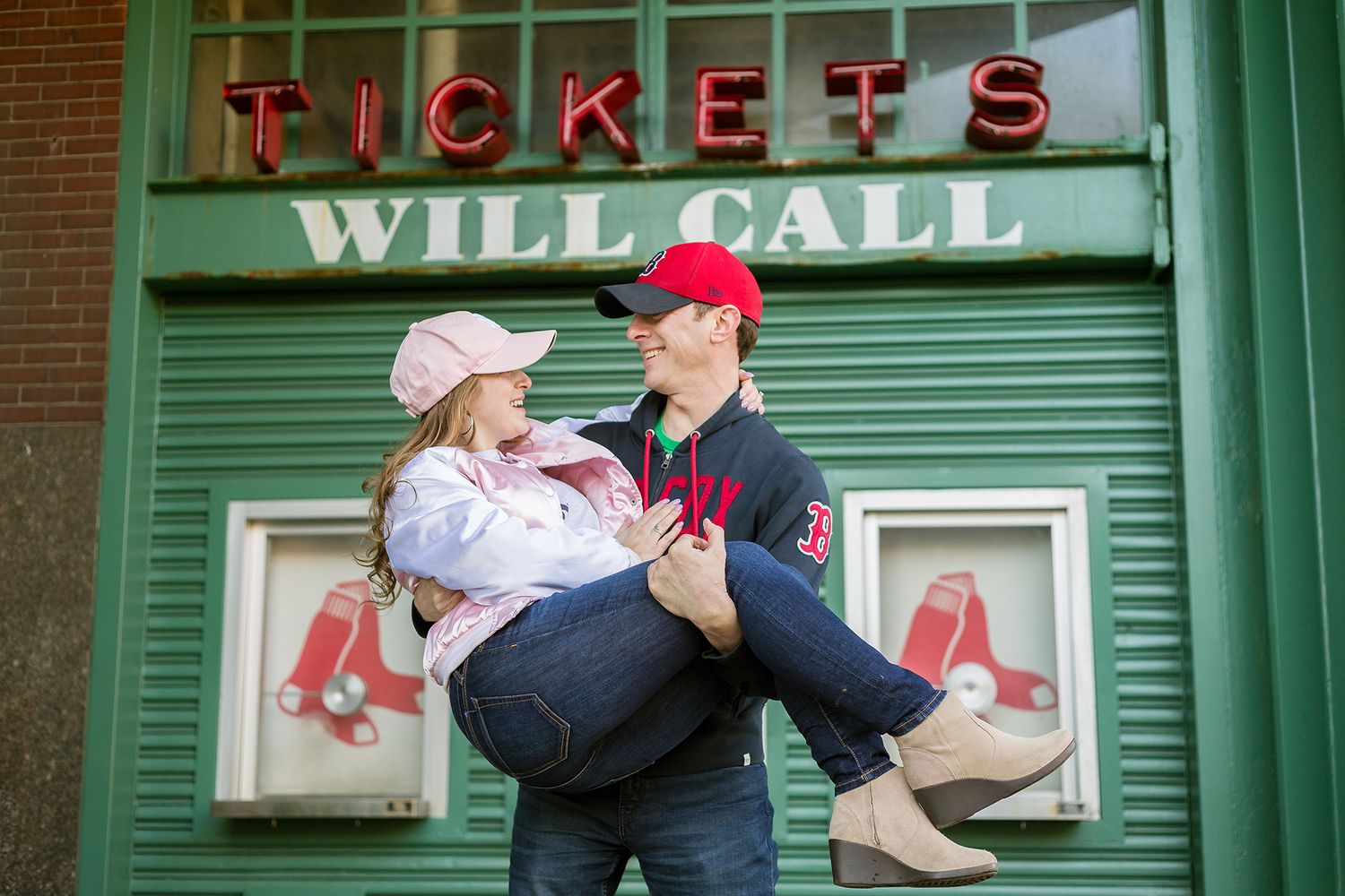 During their engagement session the bride and groom share a funny pose in front of the box office at Fenway park