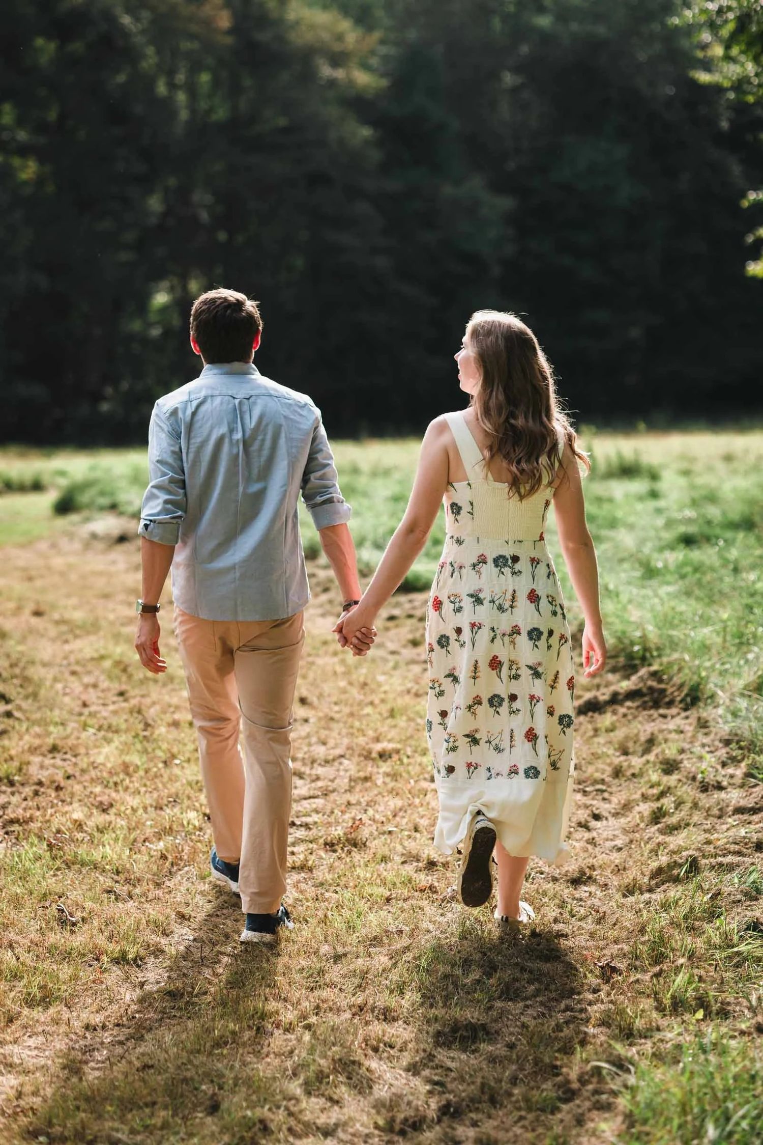 Couple holding hands walking through a sunlit meadow, woman in floral dress, man in blue shirt and khaki pants.
