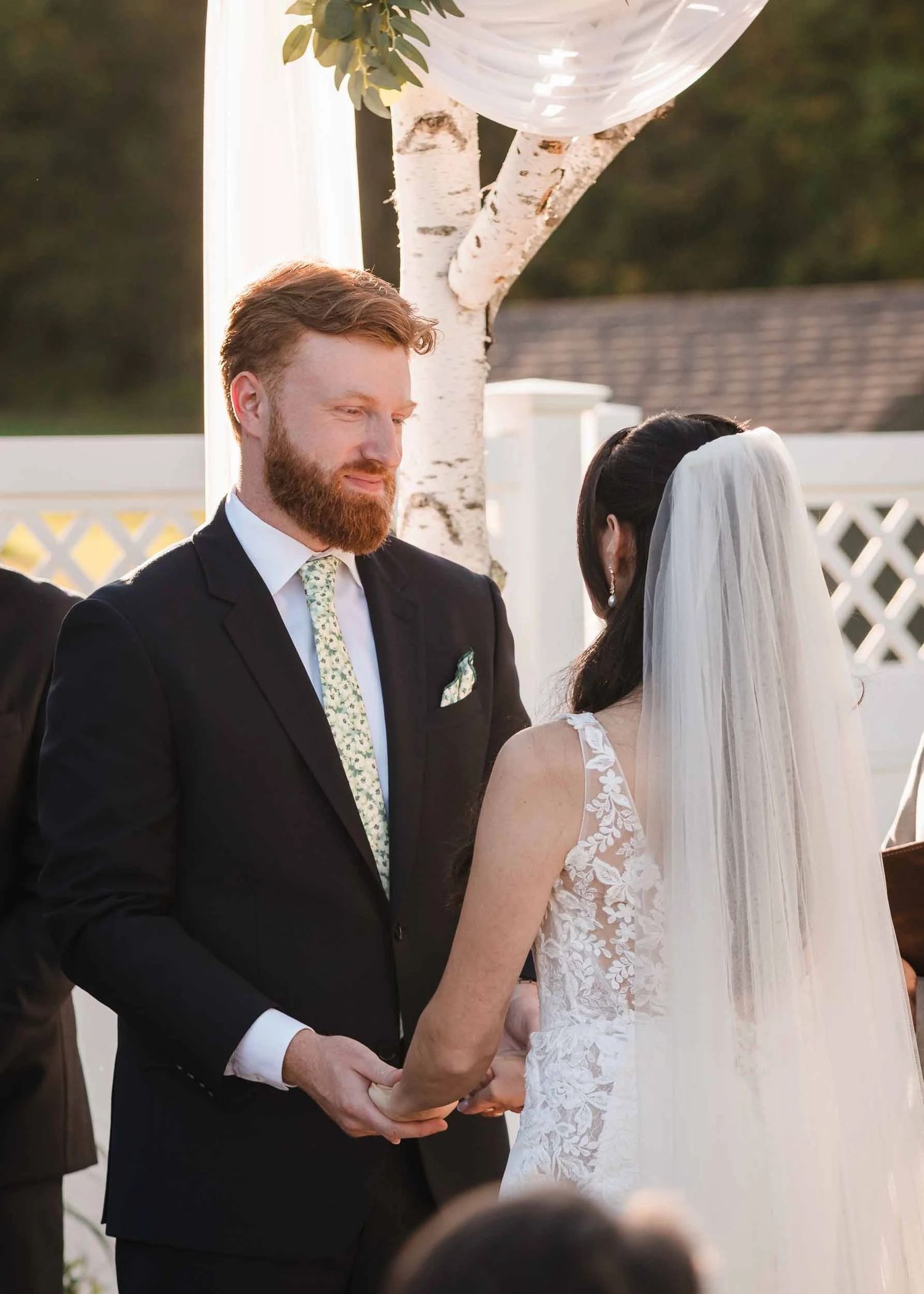 The handsome groom with red hair and a beard smiles subtly at his soon to be wife while exchanging vows during their summer wedding ceremony