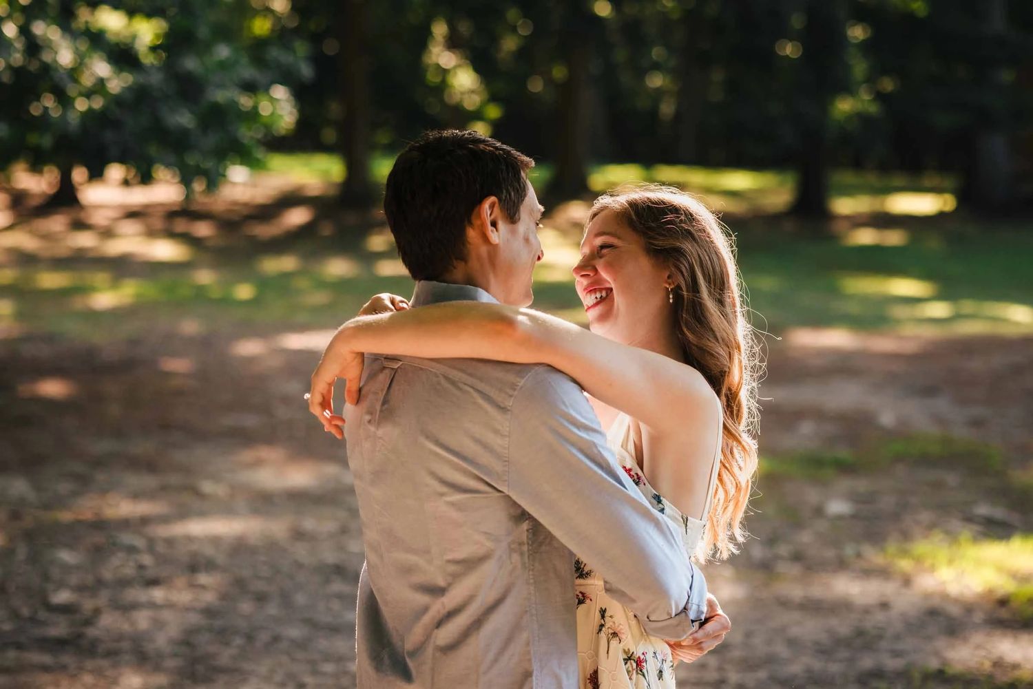 Young couple embracing and laughing together in a sunlit park path. Engagement at Borderland State Park, North Easton, MA