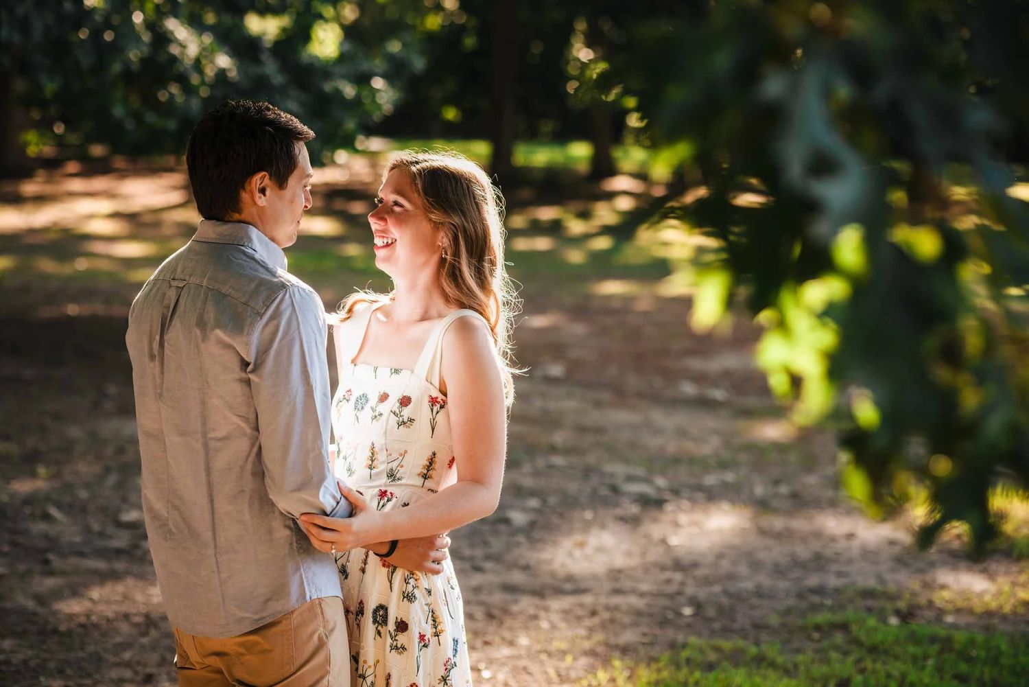 Couple standing face-to-face on a tree-lined path, smiling at each other in an intimate moment. Engagement at Borderland State Park, North