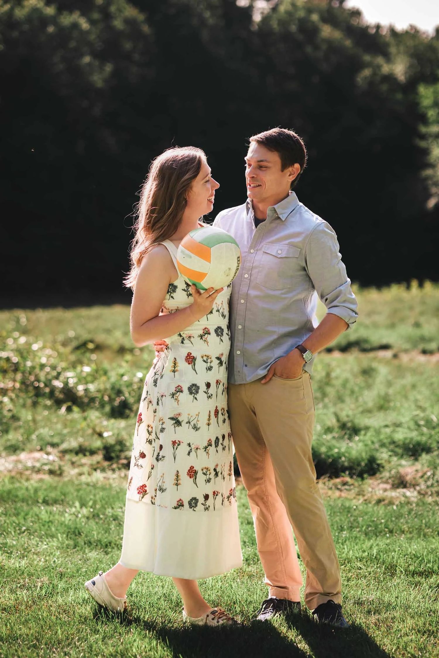Couple standing together in a sunlit field, smiling at each other while holding a volleyball Engagement at Borderland State Park