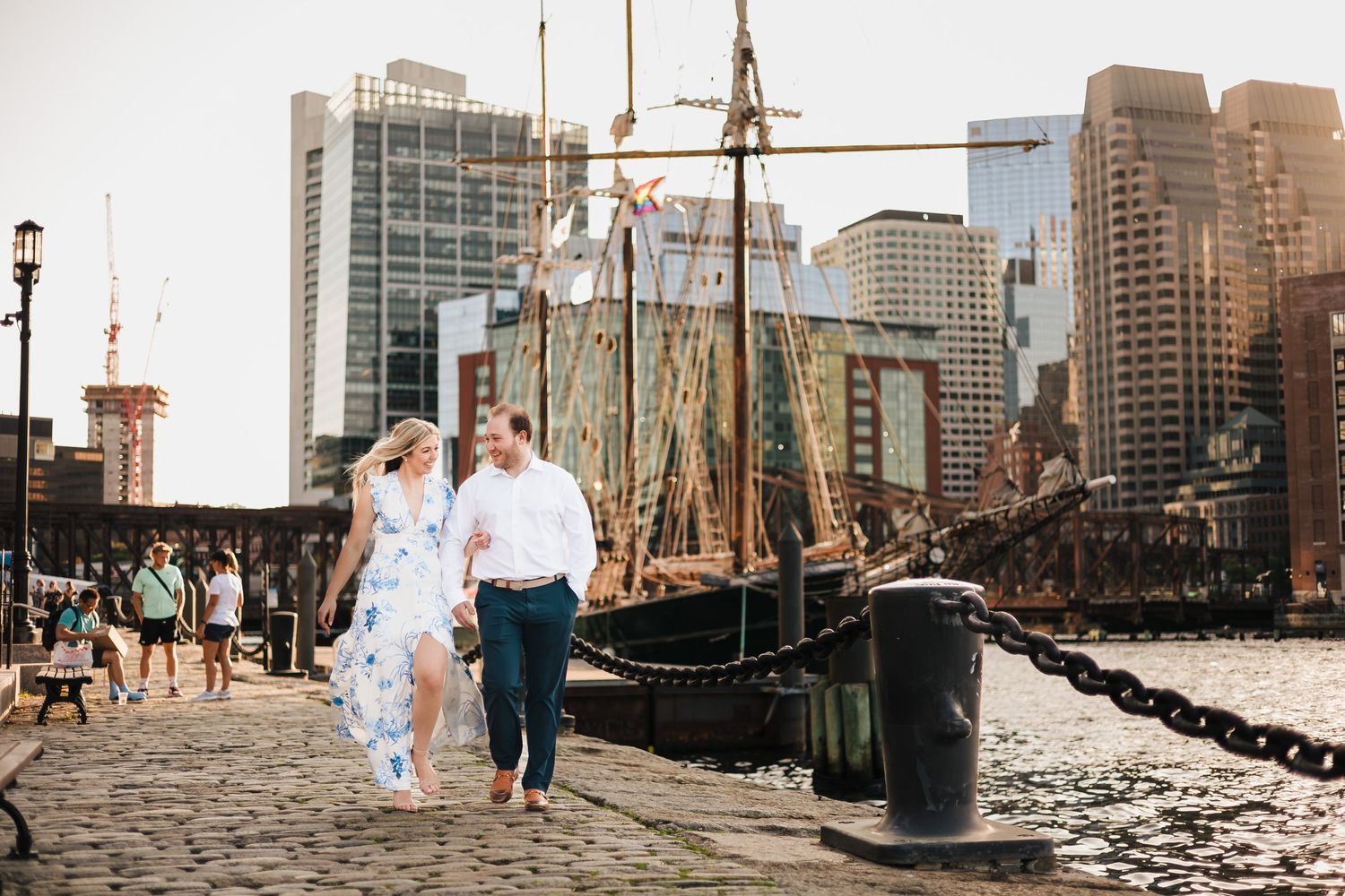 A loving couple strolls along the edge of Boston Harbor on a sunny summer day for their engagement session.
