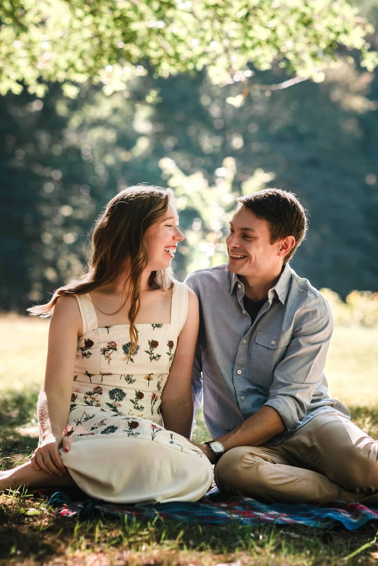 A couple sits together on a picnic blanket, smiling and gazing at each other in dappled sunlight. Engagement at Borderland State Park