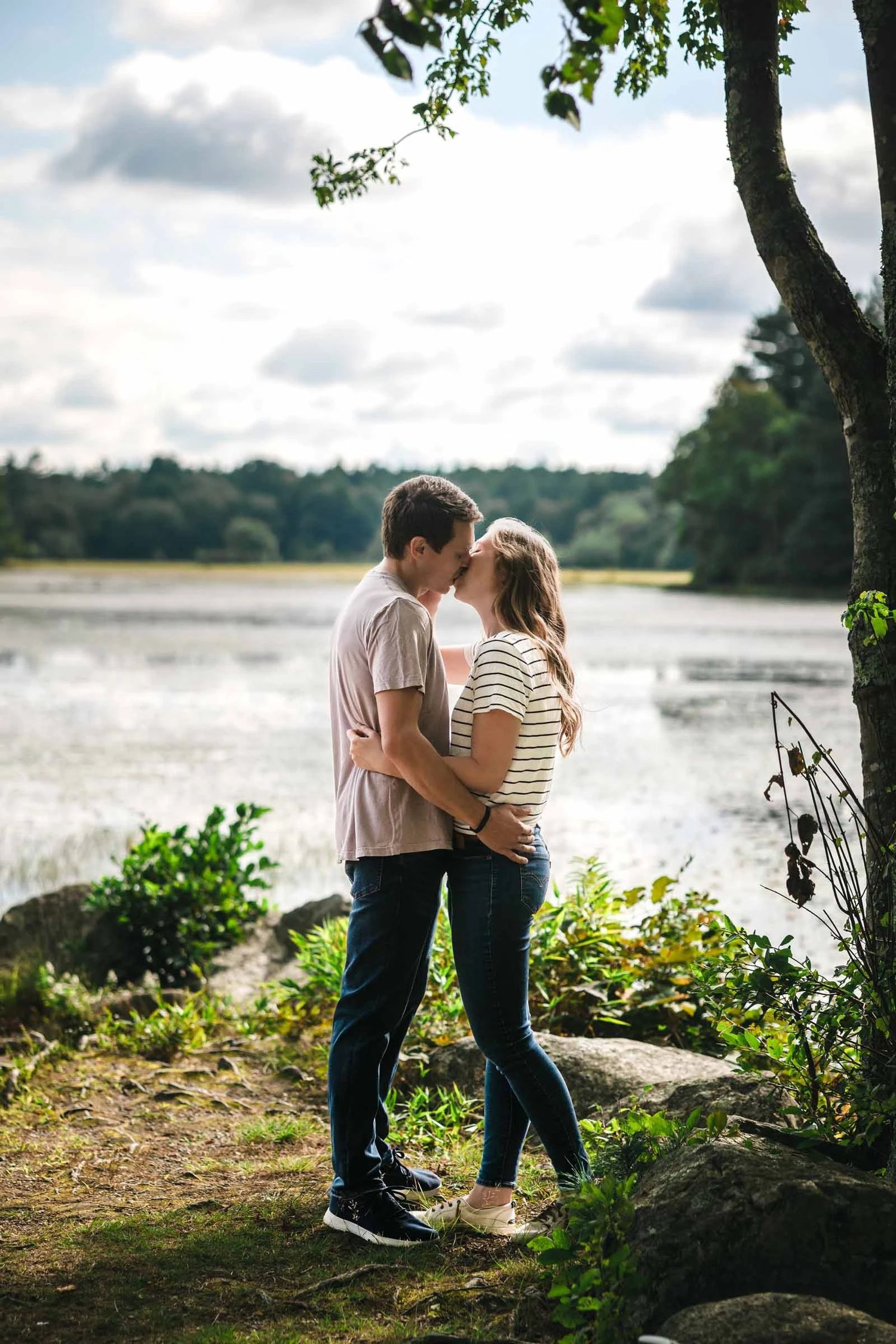 Young couple embracing by a riverside, standing beneath a leaning tree. Engagement at Borderland State Park, North Easton, MA