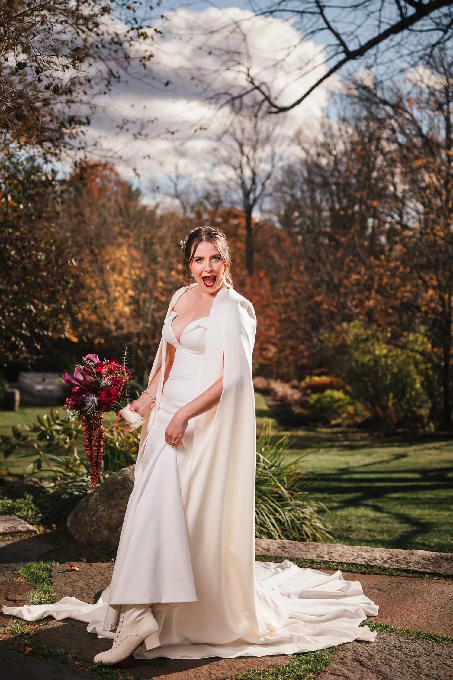 Bride standing alone in a garden, holding a deep red bouquet and gazing toward the camera for a dramatic wedding portrait
