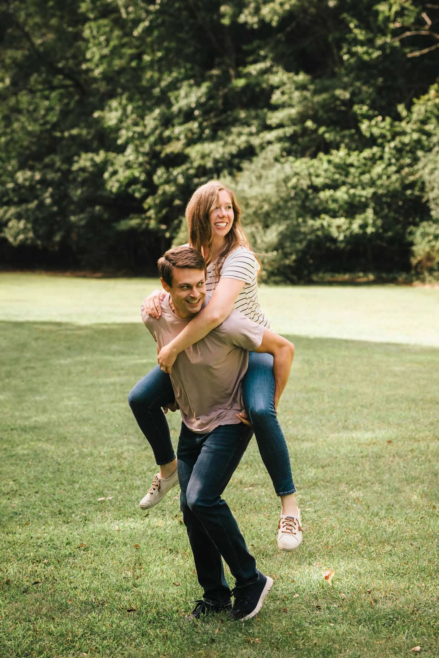 Man giving woman a piggyback ride across a grassy lawn, both laughing. Engagement at Borderland State Park, North Easton, MA