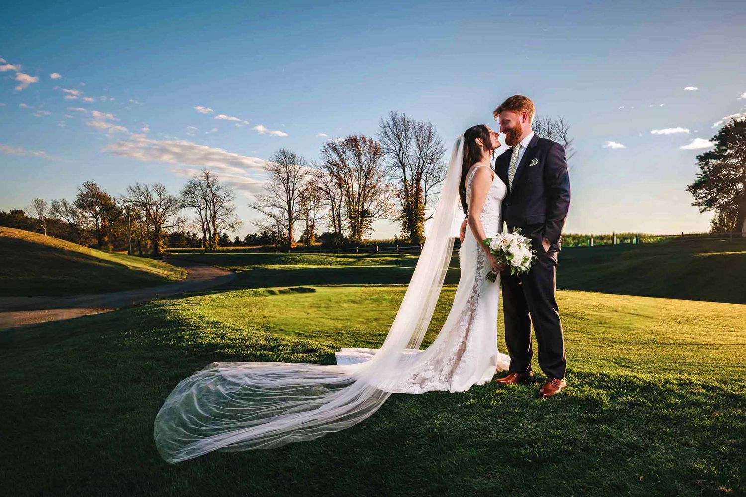 Bride and groom share a romantic moment on a lush green golf course at golden hour with a flowing cathedral veil.