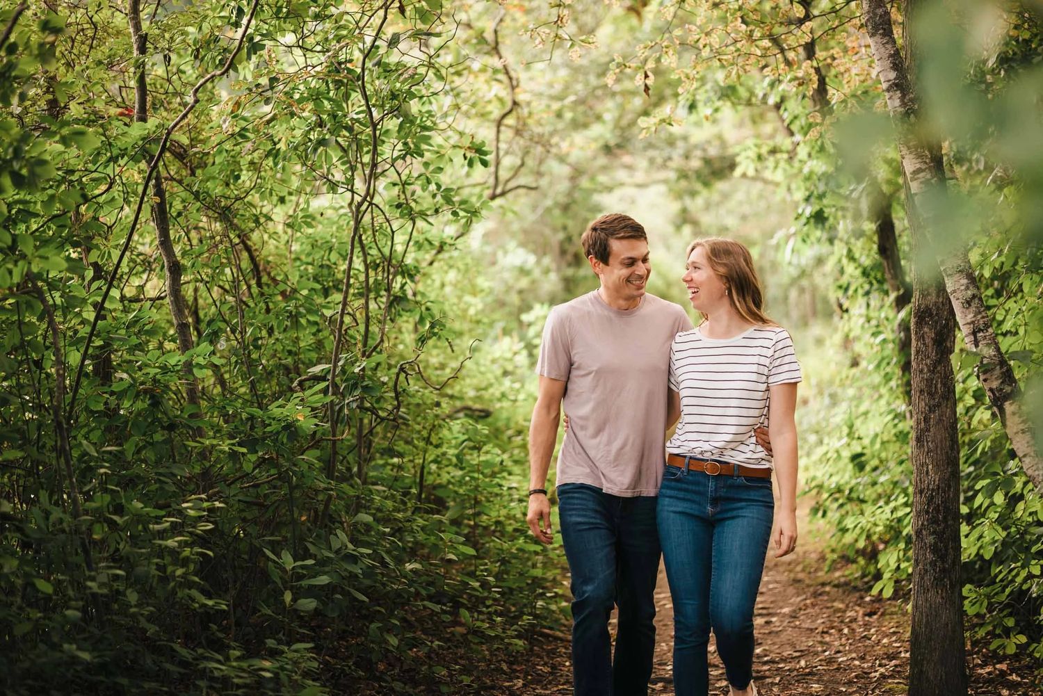 A couple walks together through a tree-lined path, looking at each other and smiling. Engagement at Borderland State Park, North Easton, M