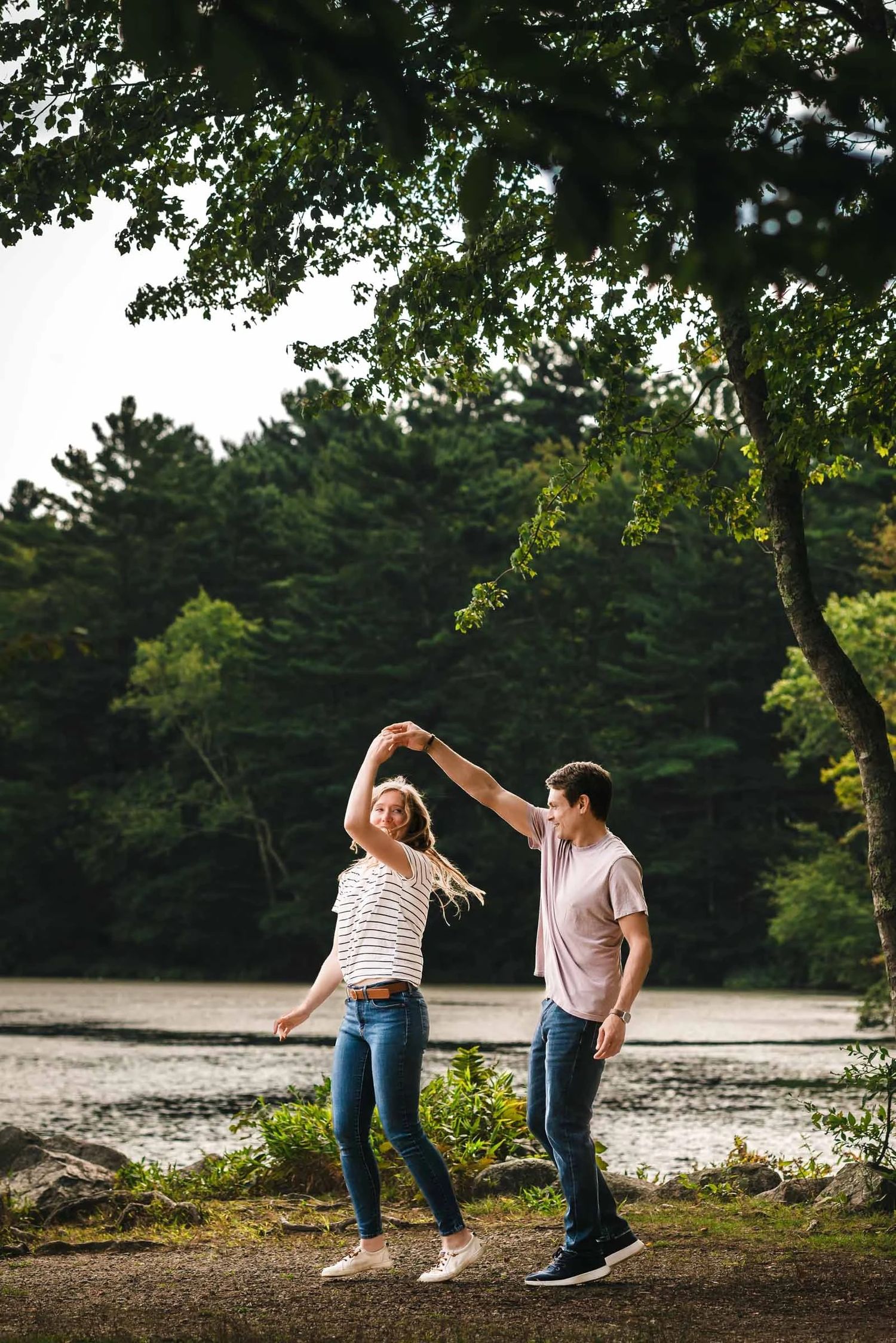 Man and woman dancing together under tree branches by a river, her arm raised overhead.  Engagement at Borderland State Park, North Easton,