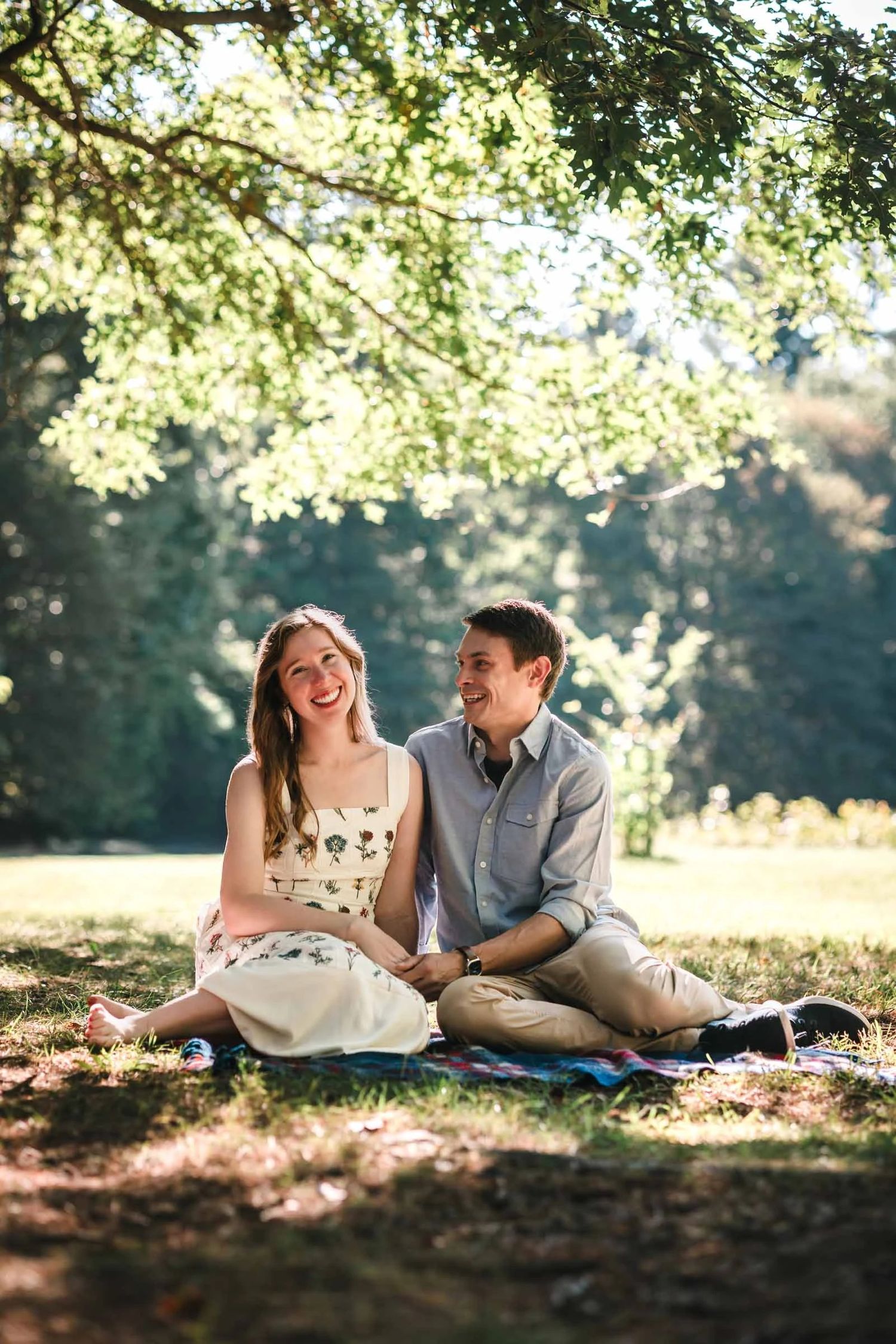 A couple sits together under dappled tree shade, gazing at each other with gentle affection. Engagement at Borderland State Park
