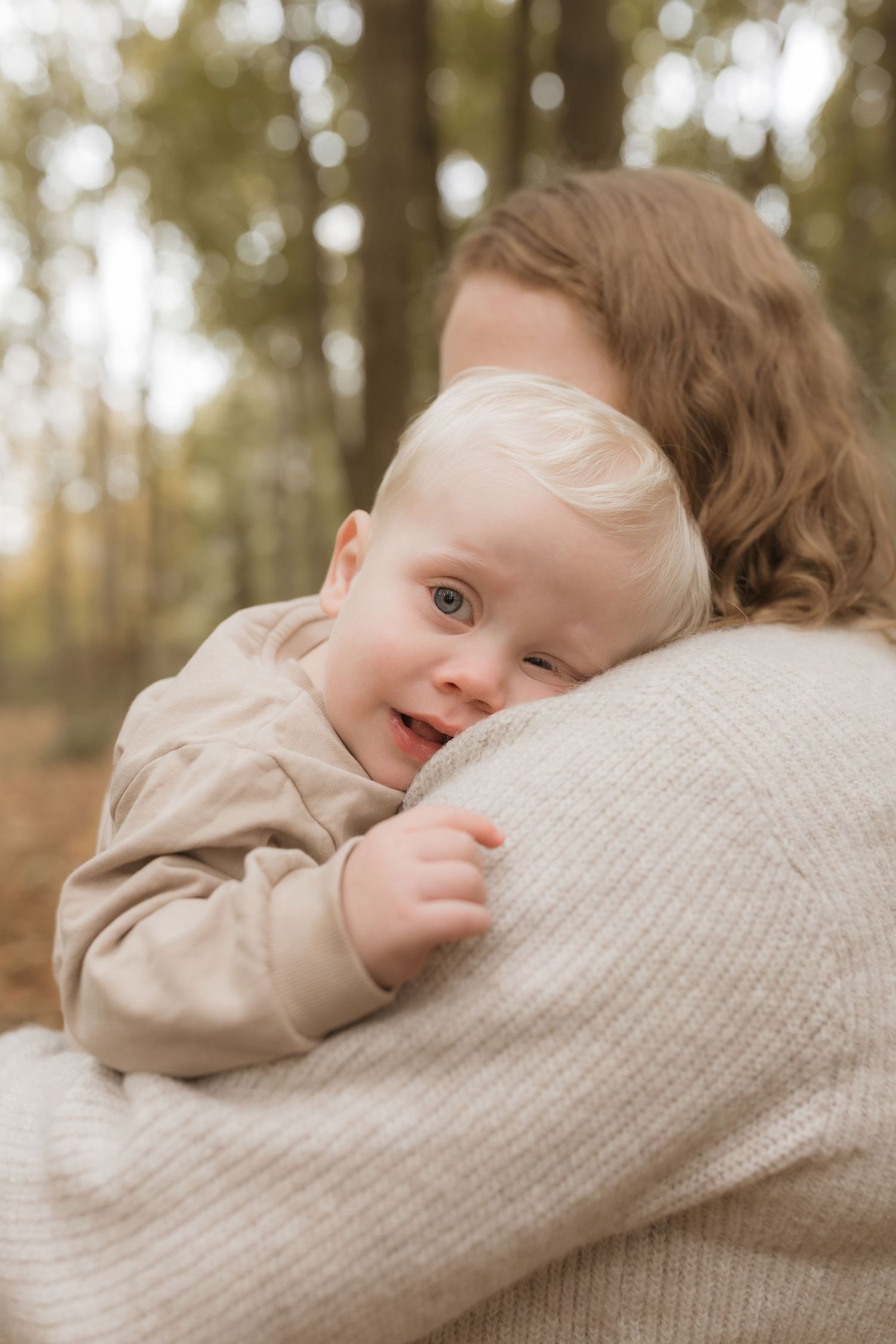 Baby with blonde hair peeks over shoulder while being held in cozy cream sweater outdoors in autumn forest setting.