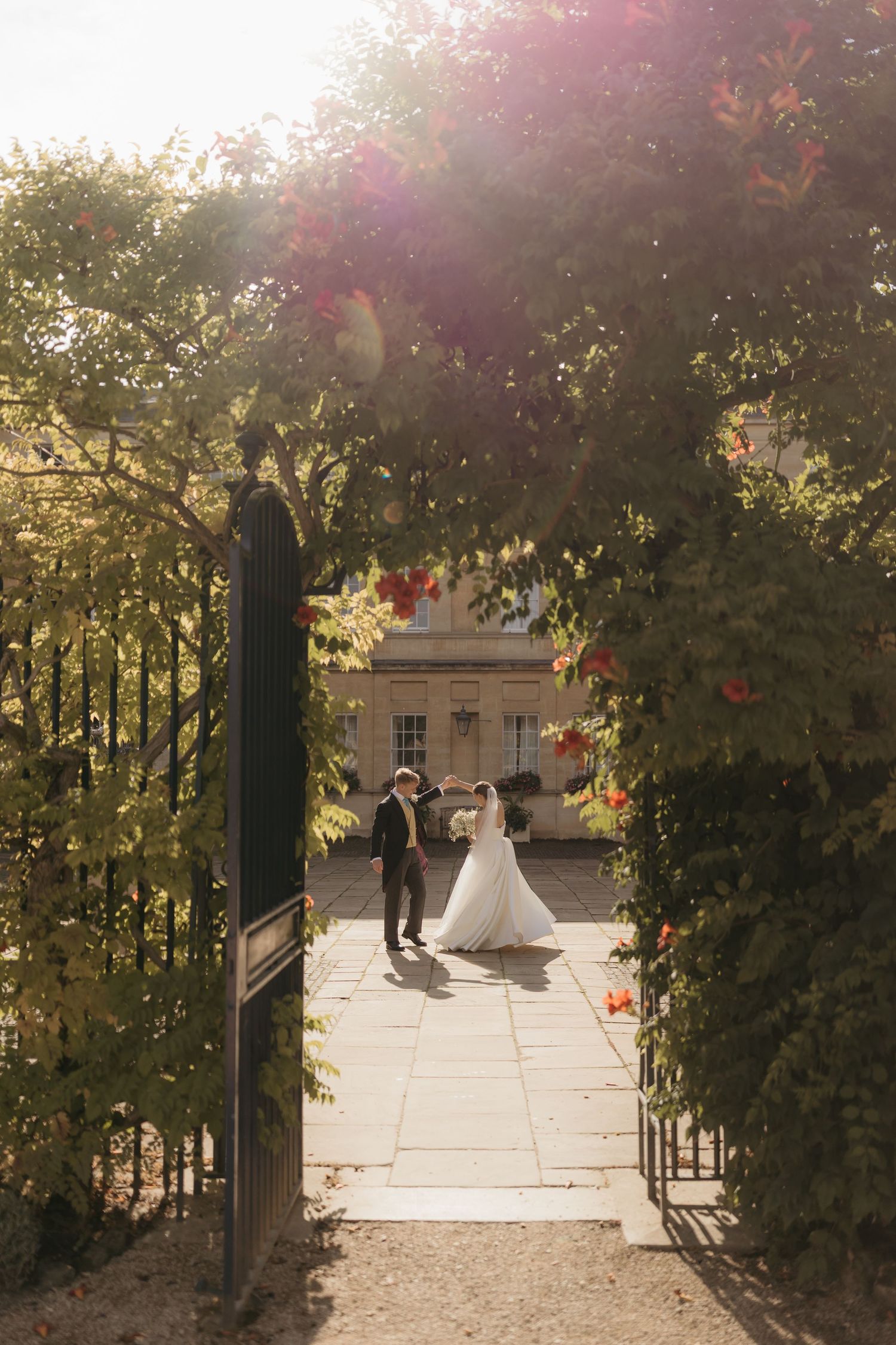 Bride and groom dancing in sunlit garden courtyard framed by iron gates and lush flowering vines.