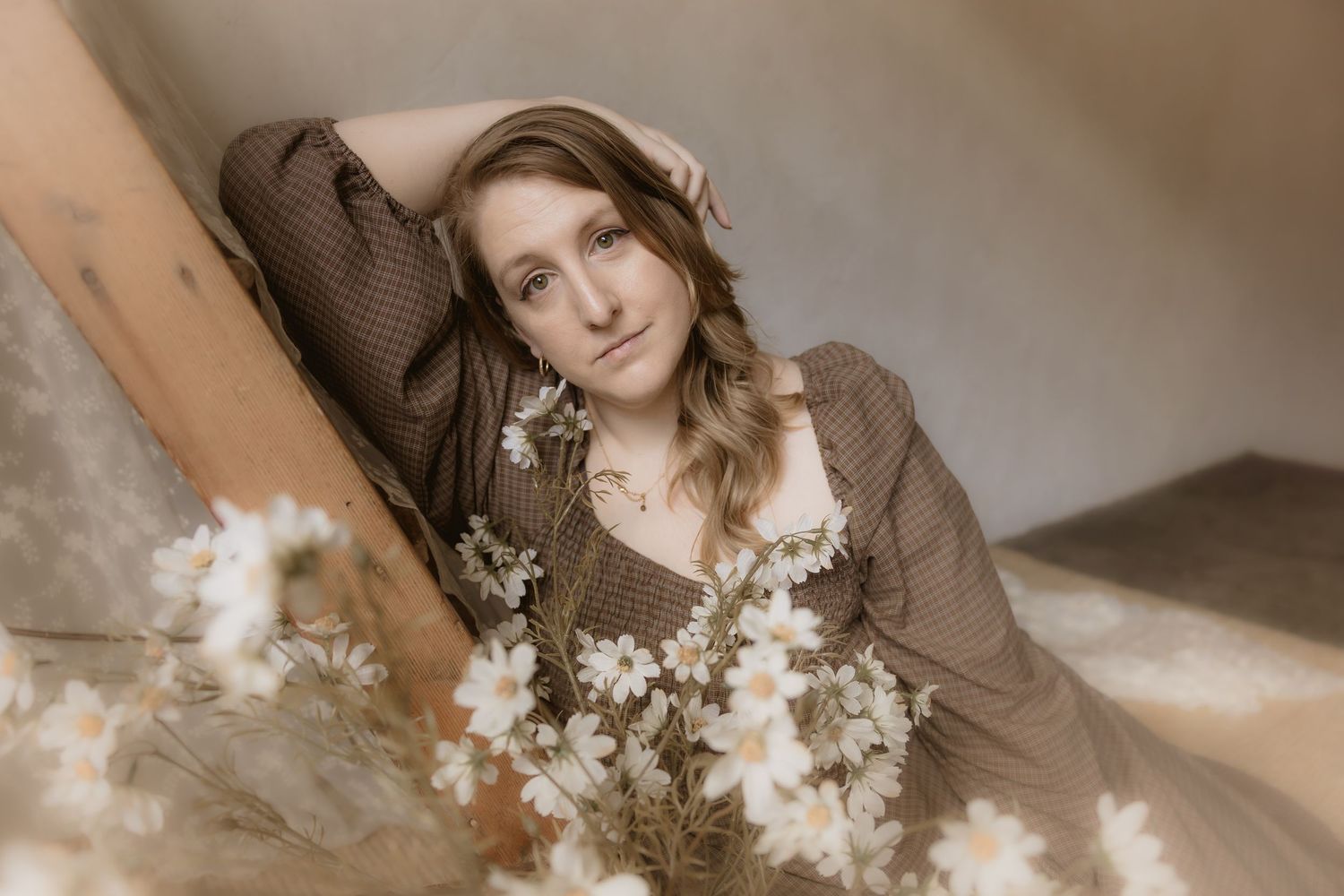 A person in a brown sweater poses with white flowers in a dreamy, ethereal photoshoot setting.