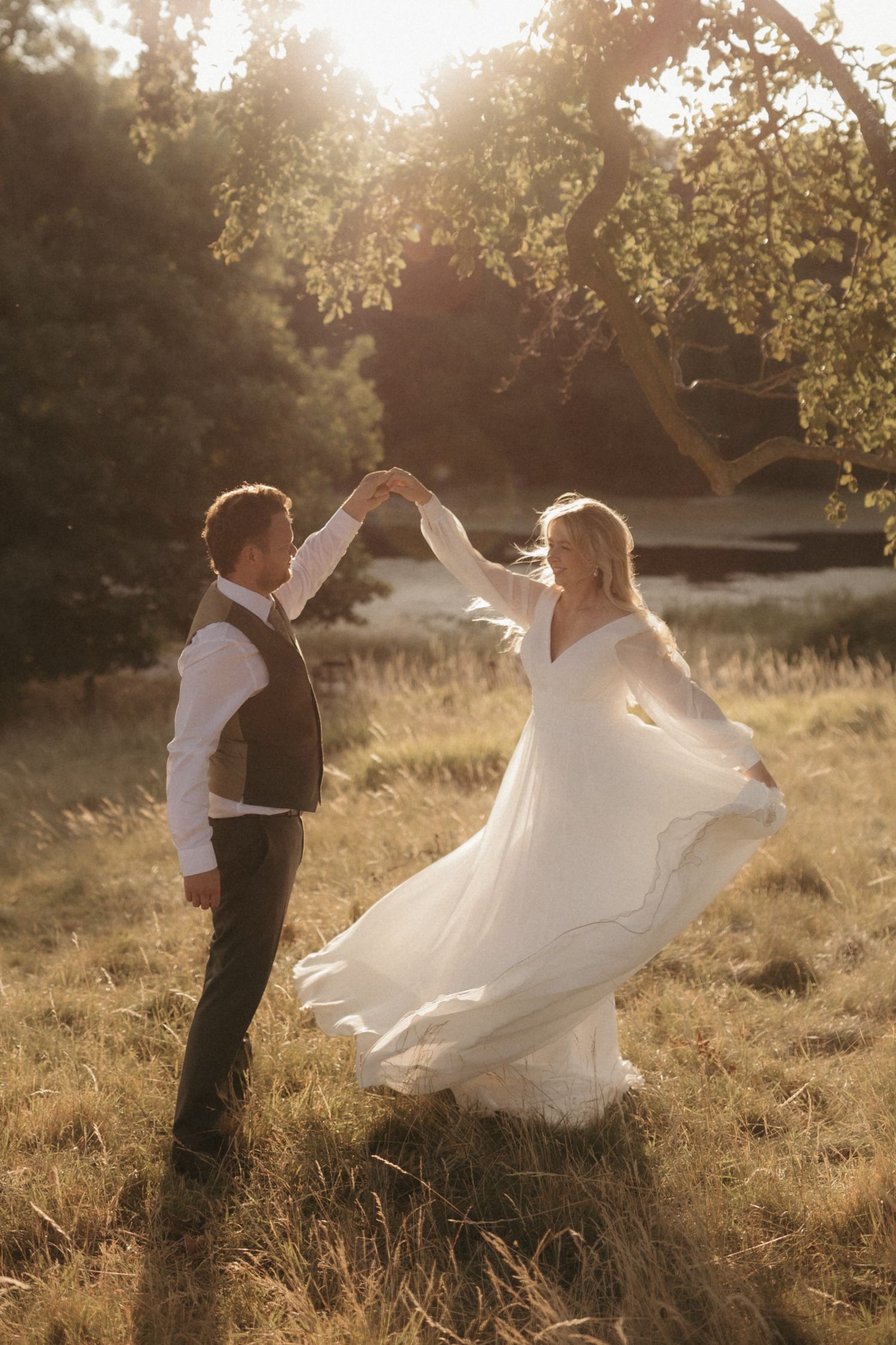 A romantic couple in wedding attire dances together in a sunlit field during golden hour.