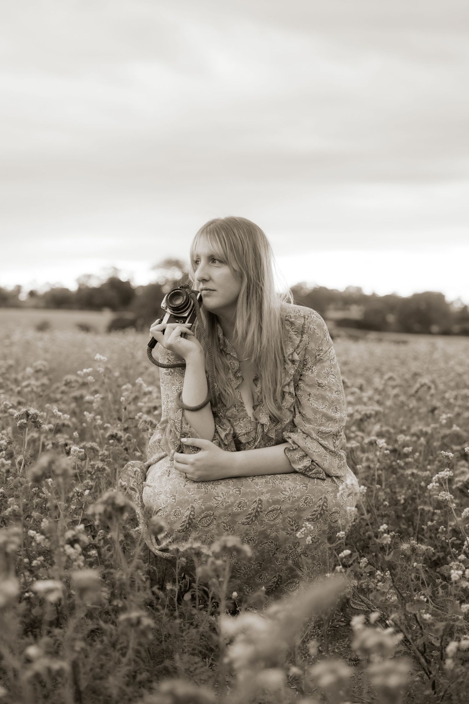 Young blonde woman in floral dress holding vintage camera in wildflower field, sepia-toned photography.