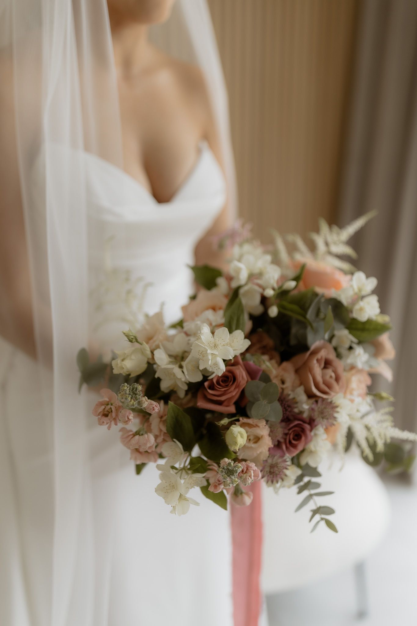 A romantic bridal bouquet with dusty rose, blush pink flowers and eucalyptus, tied with a pink ribbon against white dress.