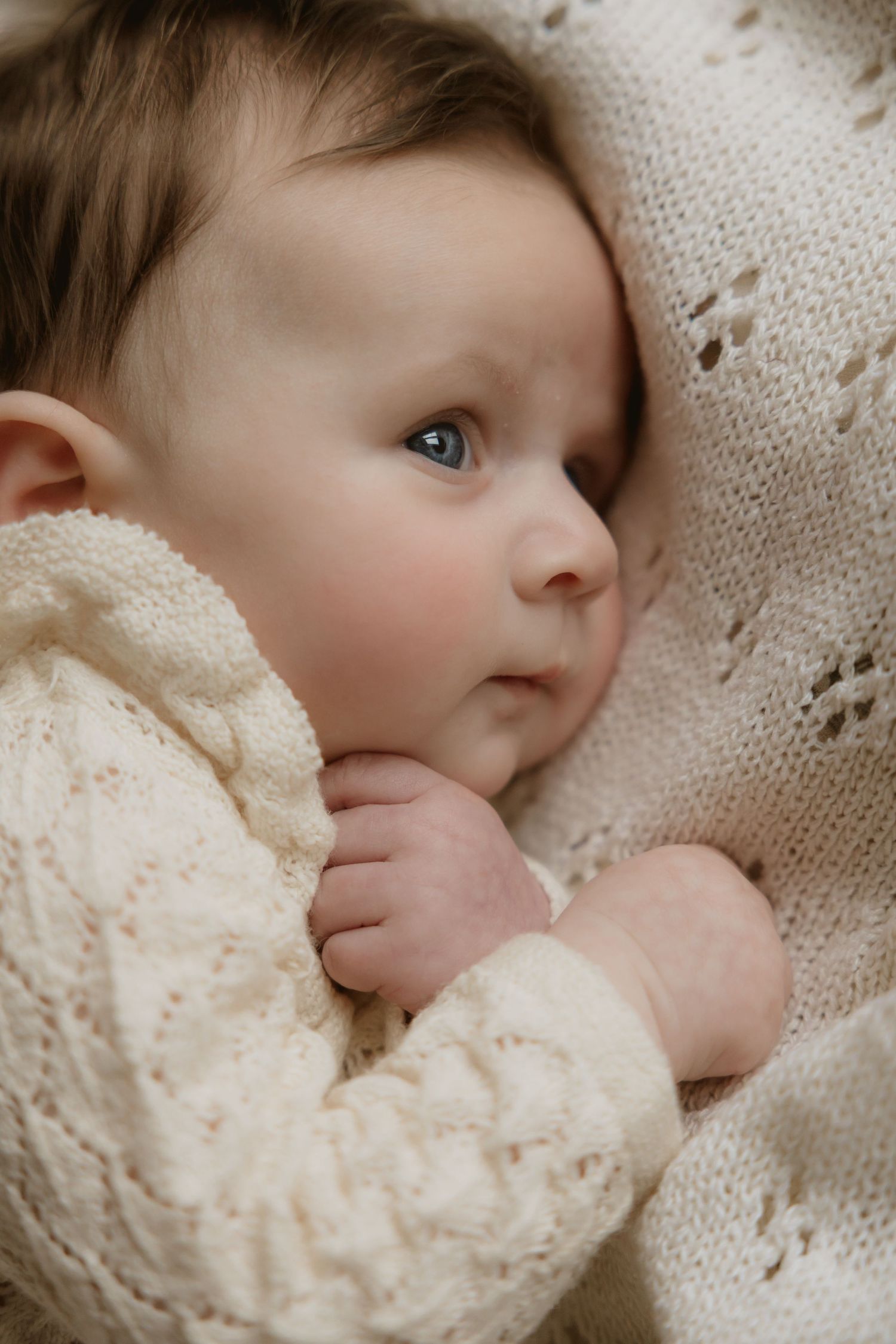 A baby in a cream knit sweater rests on soft white bedding while looking up with bright blue eyes.