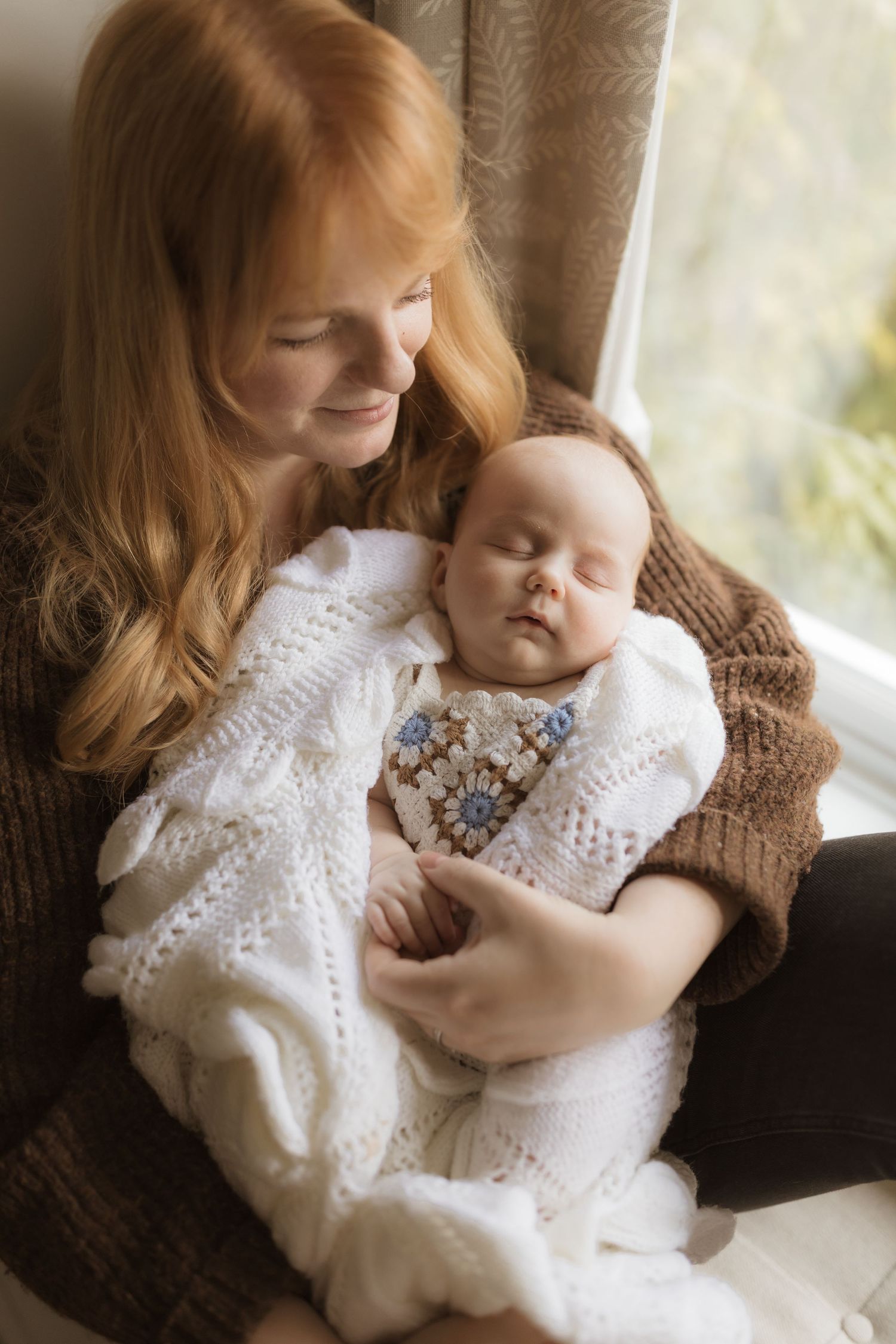 A parent in a white dress holds and comforts a sleeping baby wearing white lace near a window.