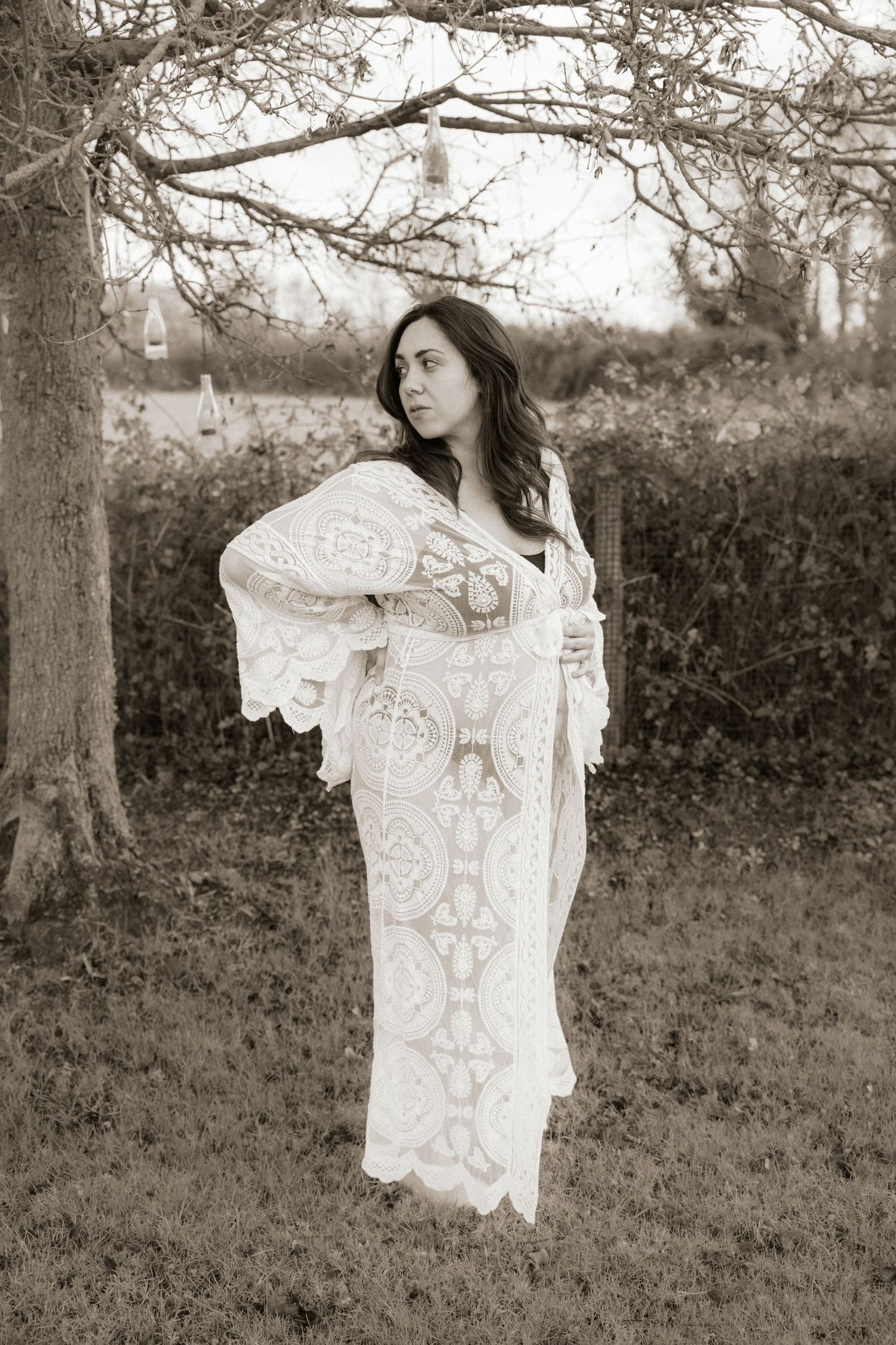 Expectant mother in flowing white lace dress posing under tree branches captured by Northamptonshire Maternity Photographer Ellen Sear
