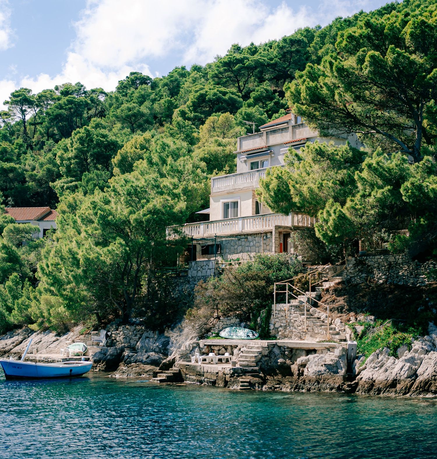 A stone villa nestled among lush pine trees on a rocky Croatian coastline with turquoise waters and a blue boat.