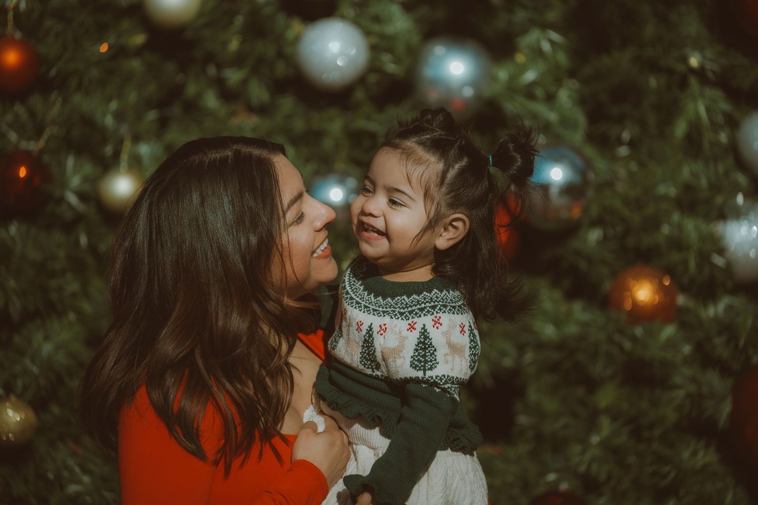 A joyful mother holds her laughing toddler daughter in front of a decorated Christmas tree.