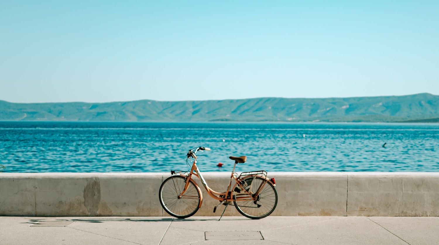 A vintage pink bicycle parked on a seaside promenade with calm blue ocean and mountains in the background.