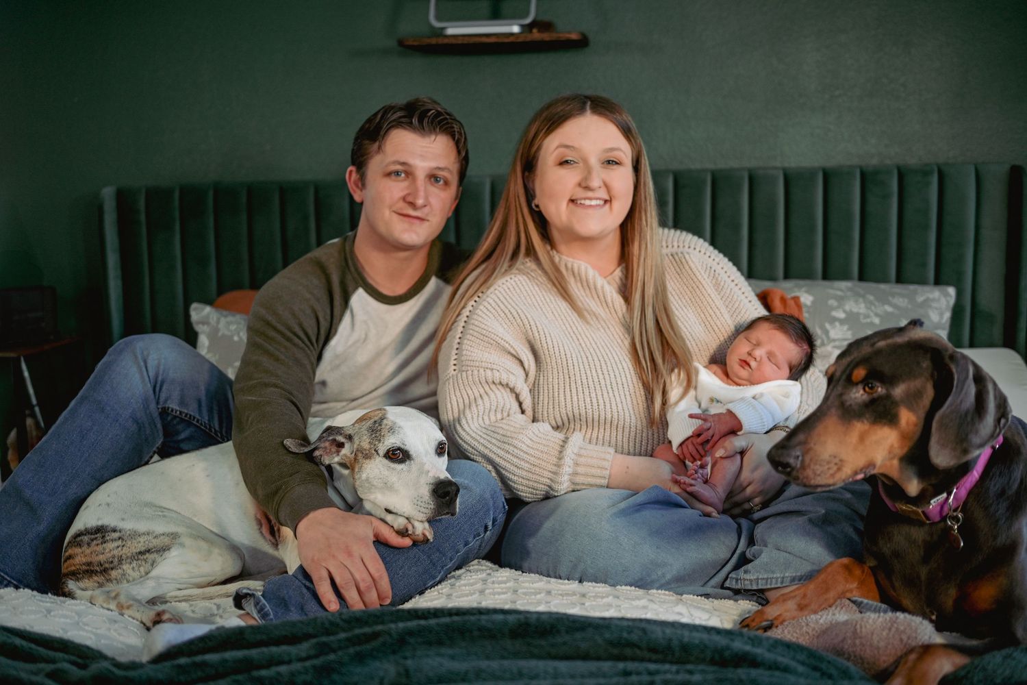 Happy couple with newborn baby and two dogs relaxing together on a green velvet bed.