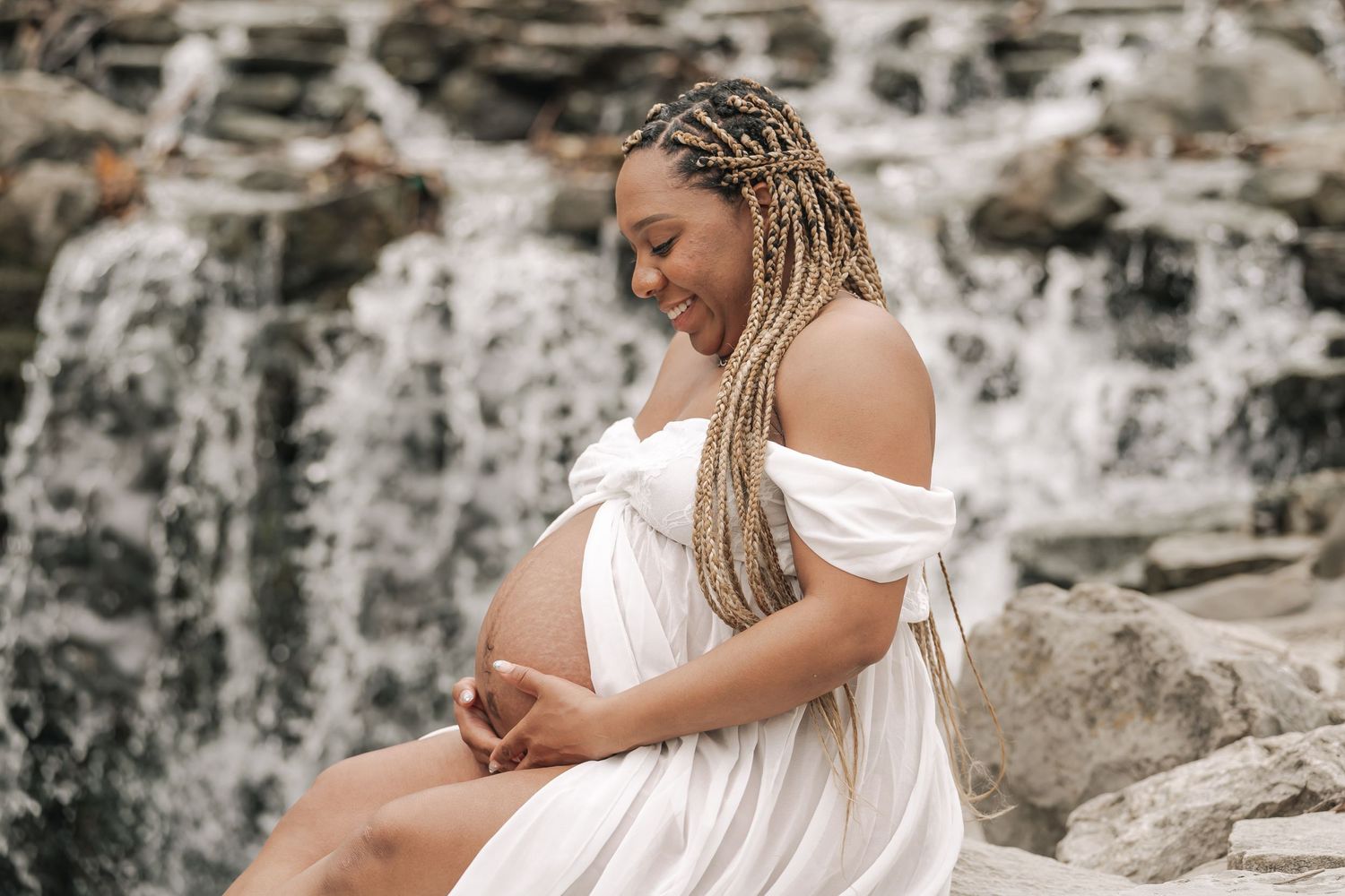 Smiling pregnant woman in white off-shoulder dress sitting by a waterfall.