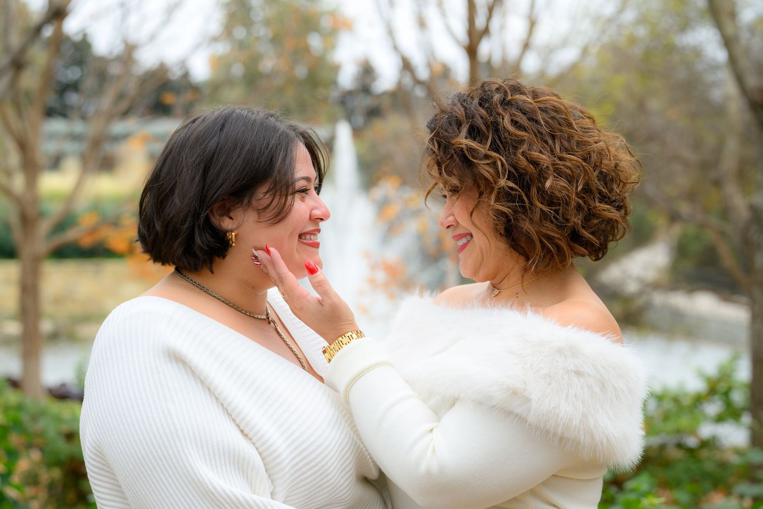 Two women in white sweaters laughing together outdoors near a fountain.