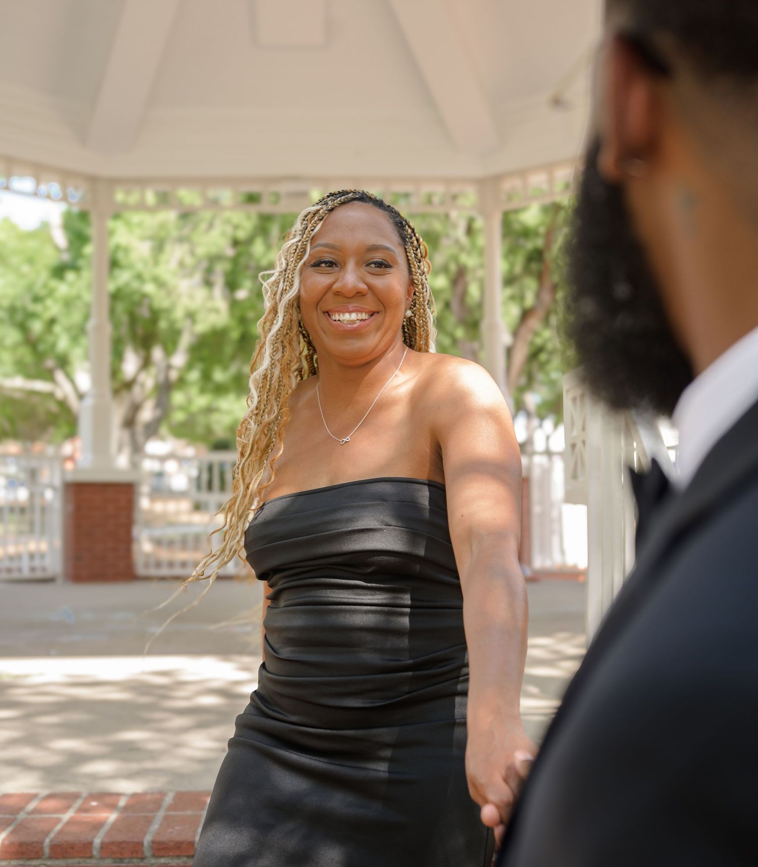 A smiling woman in a black strapless dress stands near a gazebo on a sunny day.