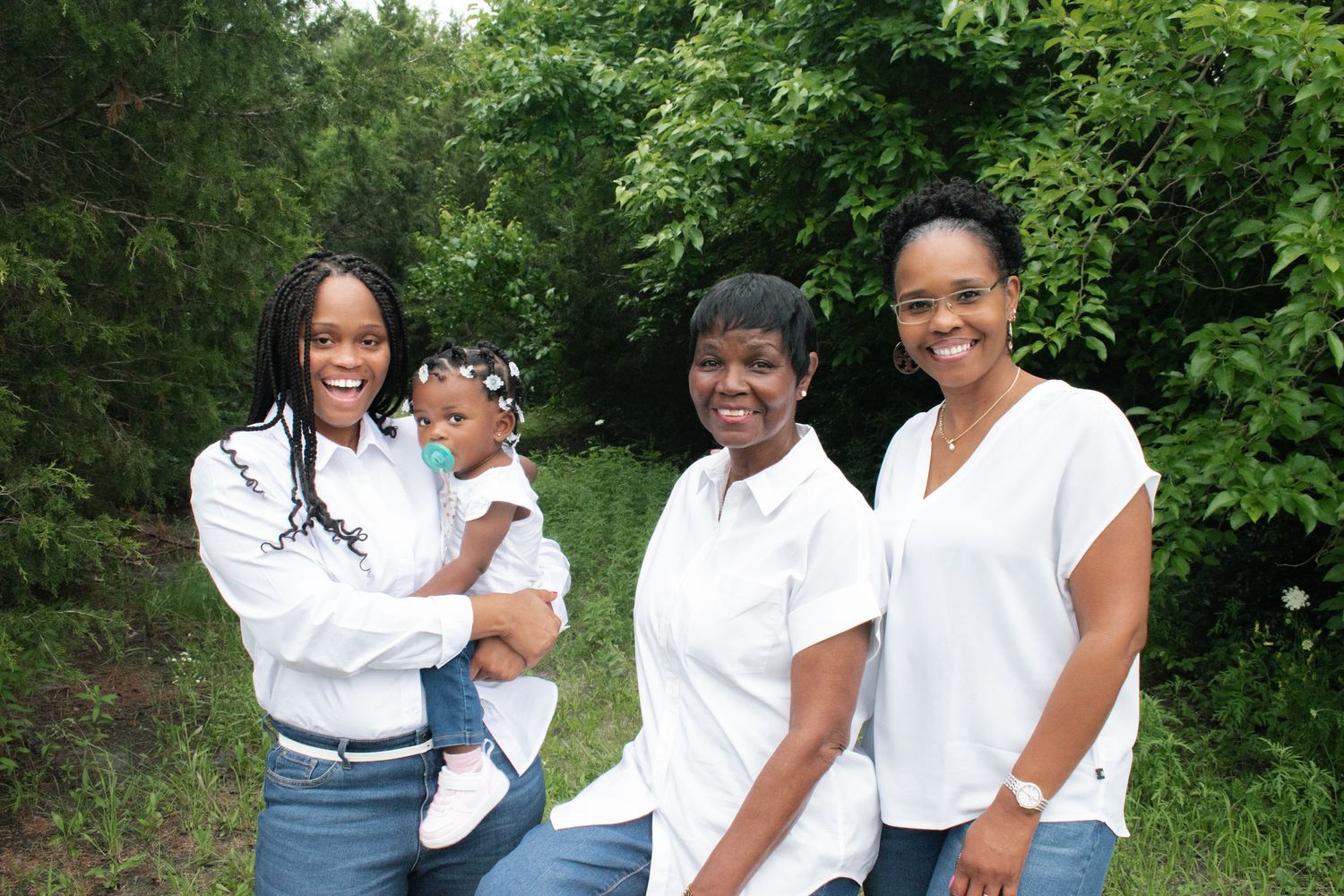 Three women in white tops smile together outdoors surrounded by lush green trees.