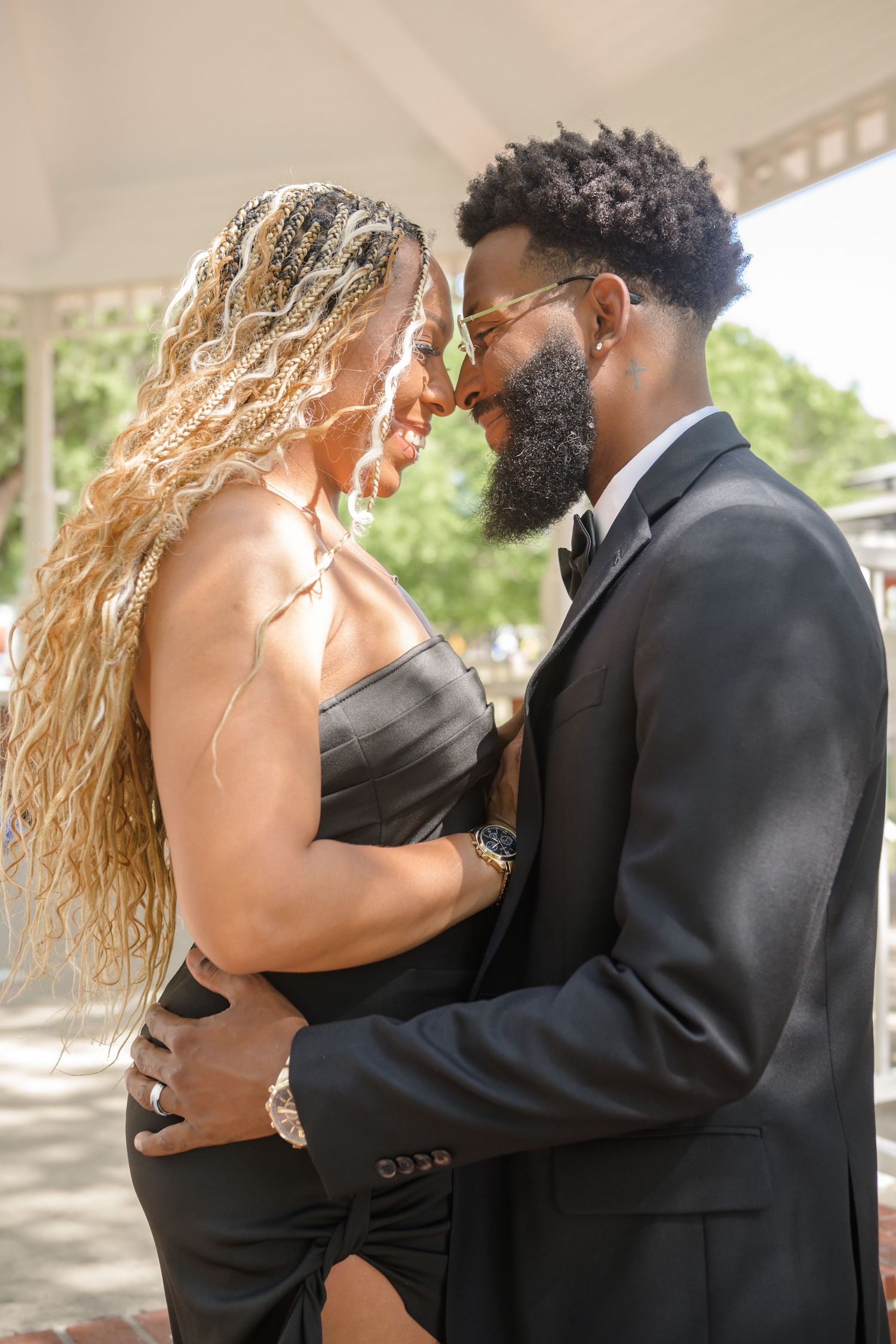 A romantic couple in black formal wear share an intimate moment under a sunny pavilion.
