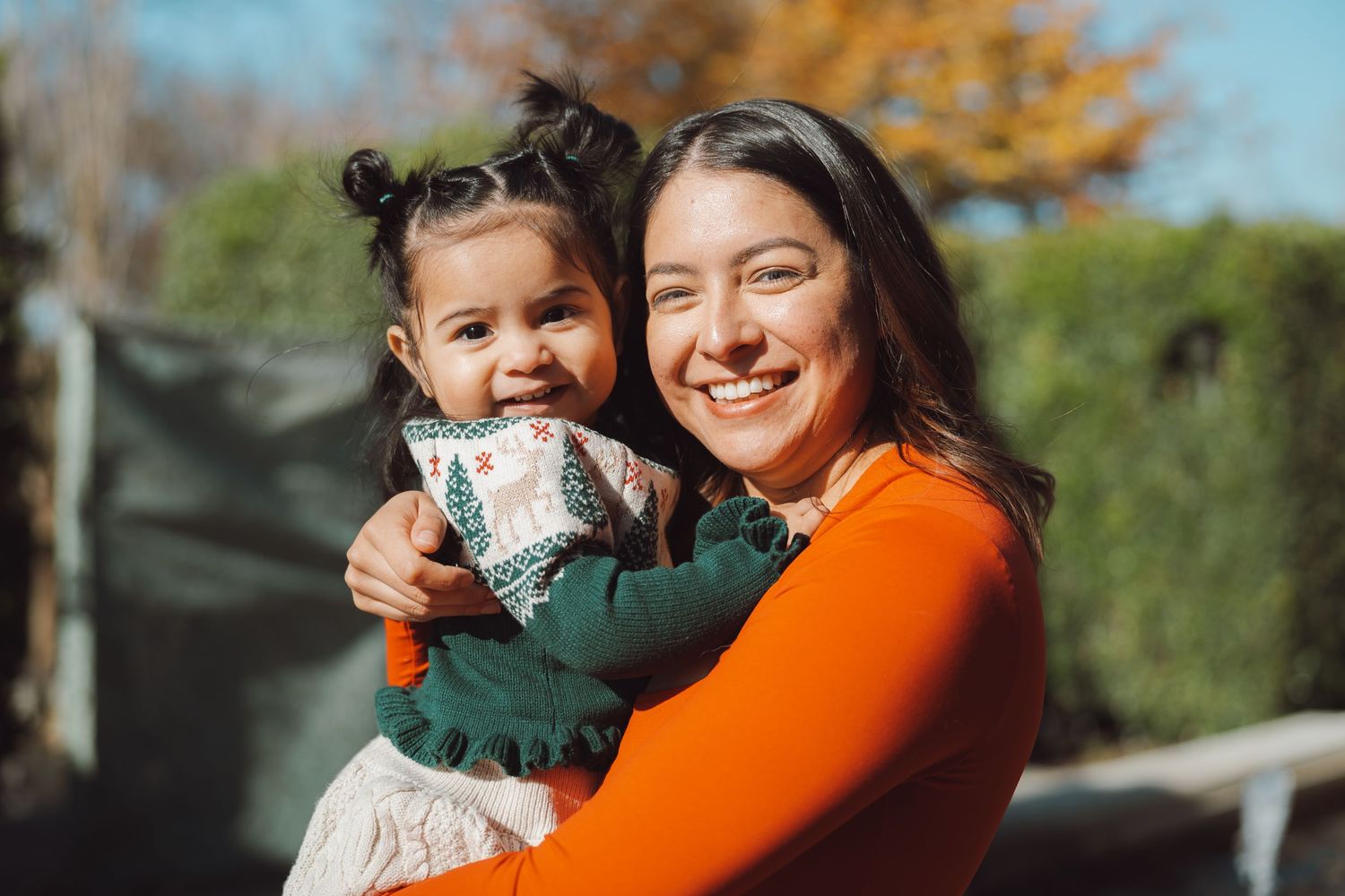 A smiling woman in an orange top holds a happy toddler girl outdoors in autumn.