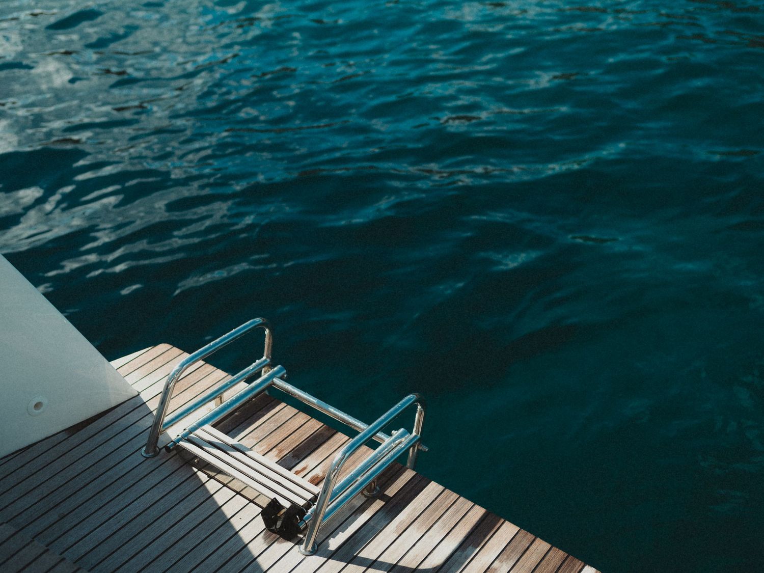 A wooden dock with metal ladder steps leading into calm turquoise blue ocean water on a sunny day.