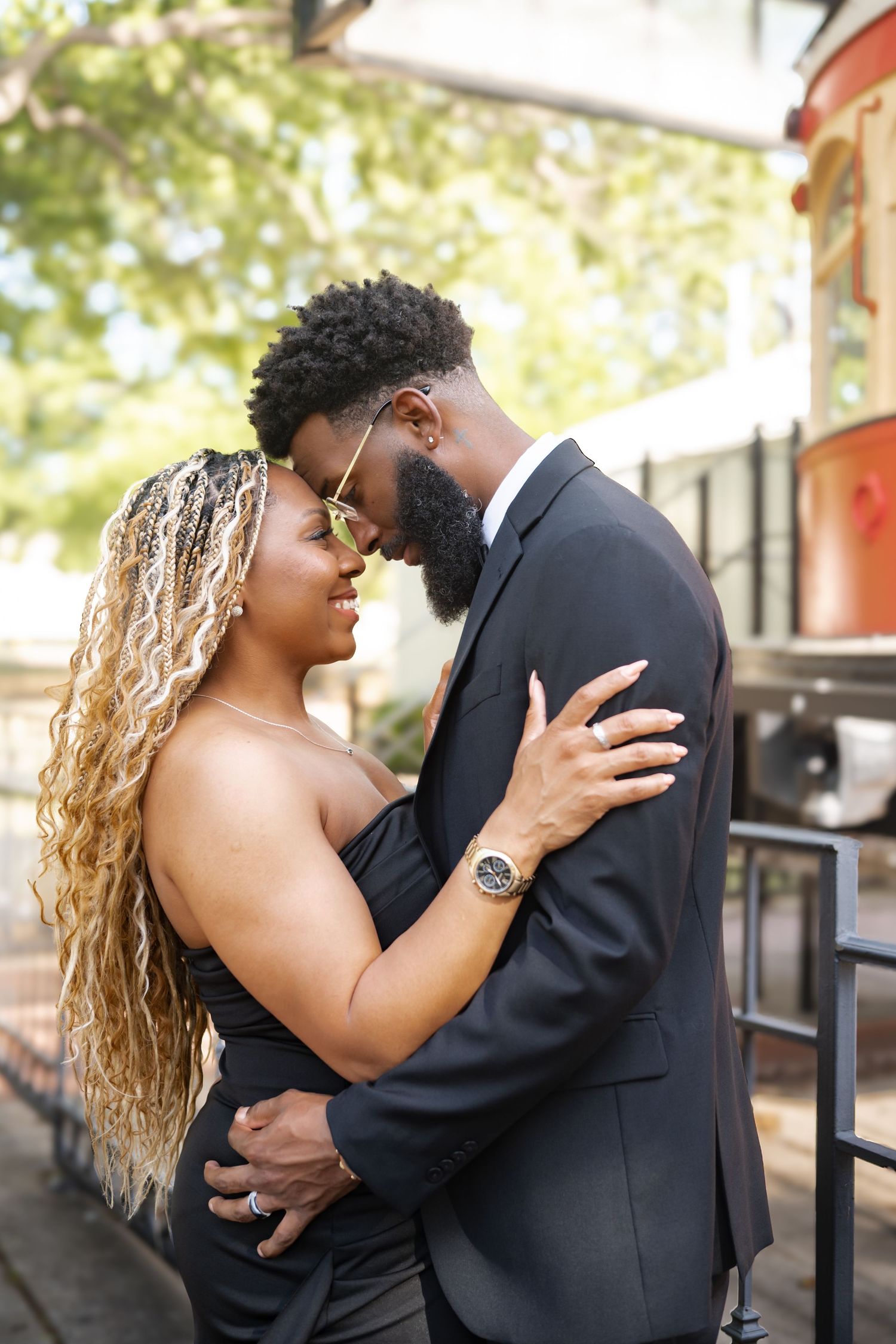 An elegant Black couple in formal black attire embrace closely outdoors on a sunny day.