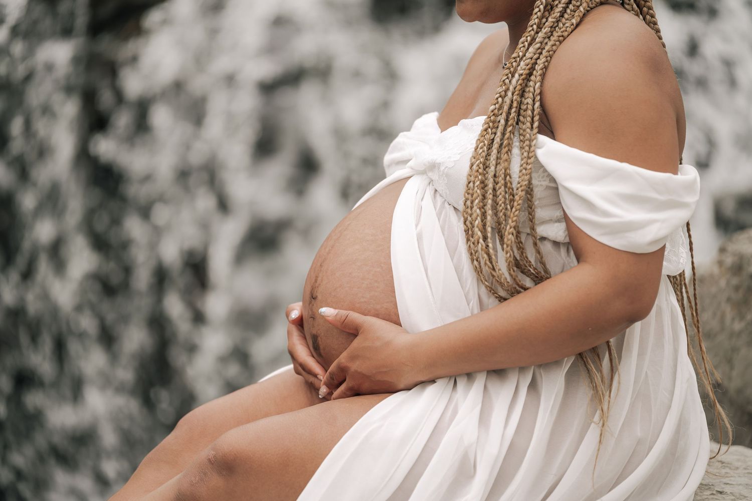 Pregnant woman in white dress cradling her baby bump while seated near a waterfall.