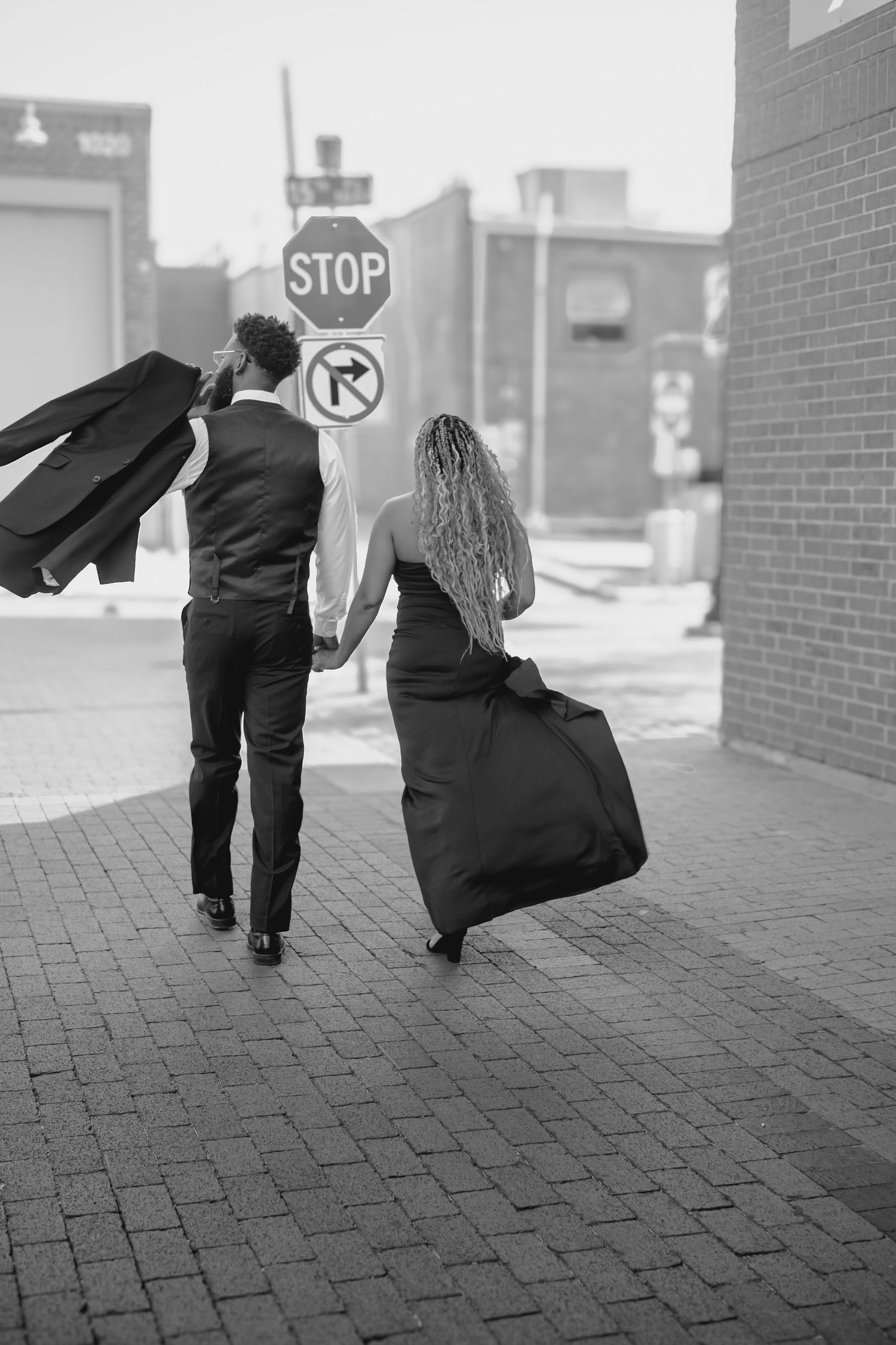 A couple in black formal attire walk hand-in-hand down a brick sidewalk in black and white.