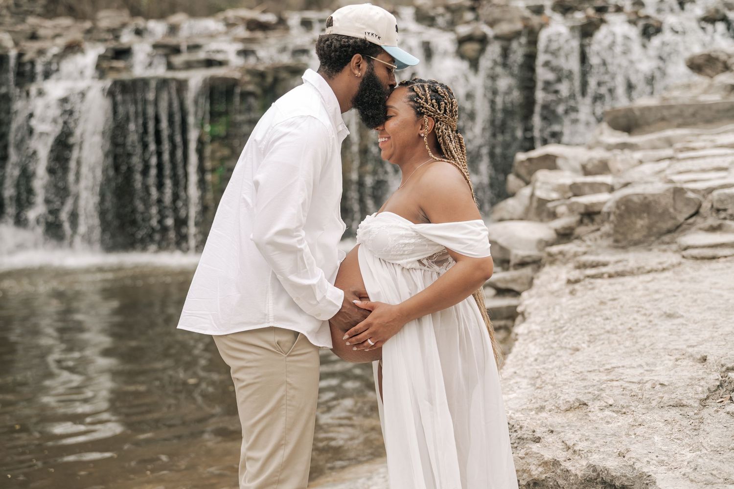 Couple in white outfits holding hands near a waterfall during a maternity photoshoot.