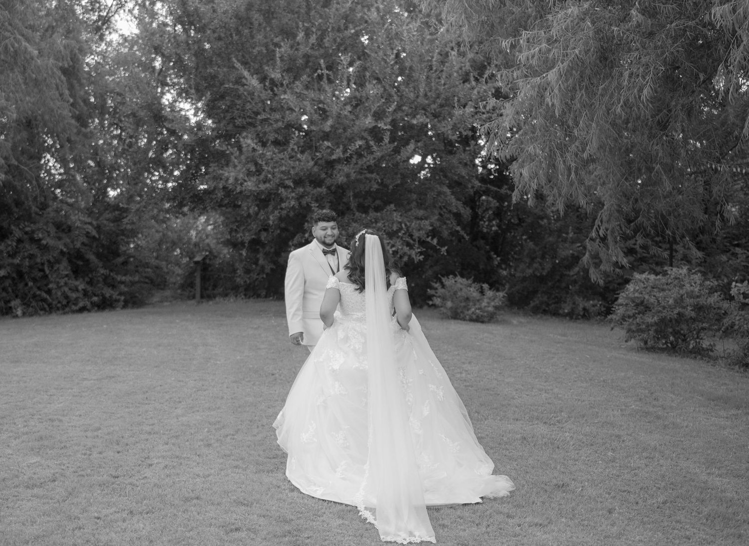 Black and white photo of a bride and groom standing together outdoors on their wedding day surrounded by lush trees.