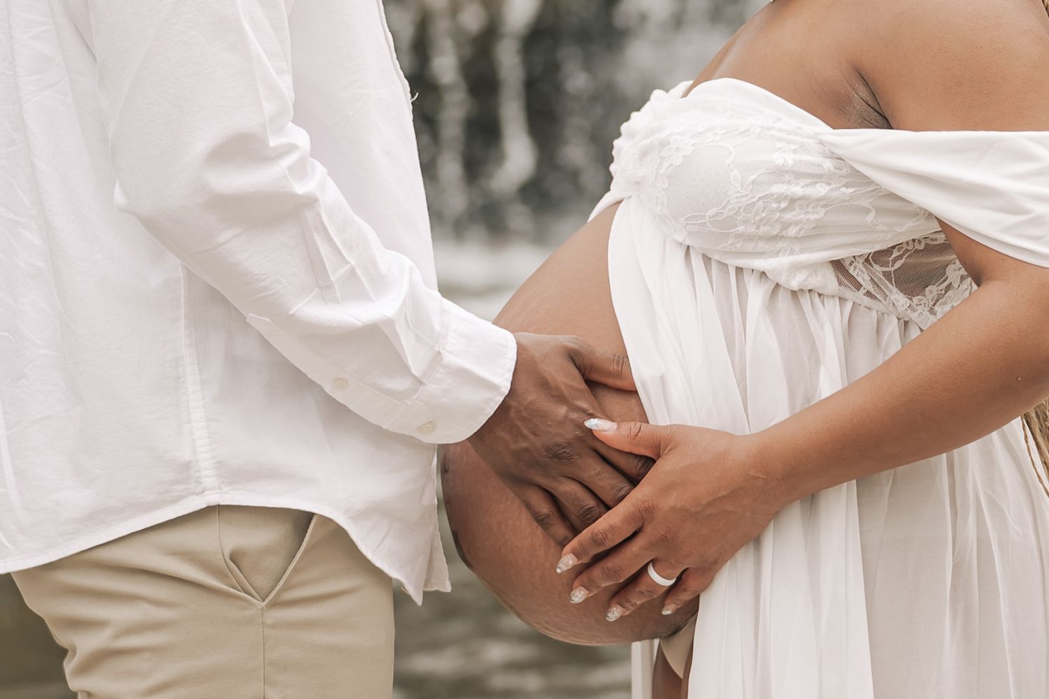 Close-up of a couple cradling a pregnant belly in white outfits near a waterfall.