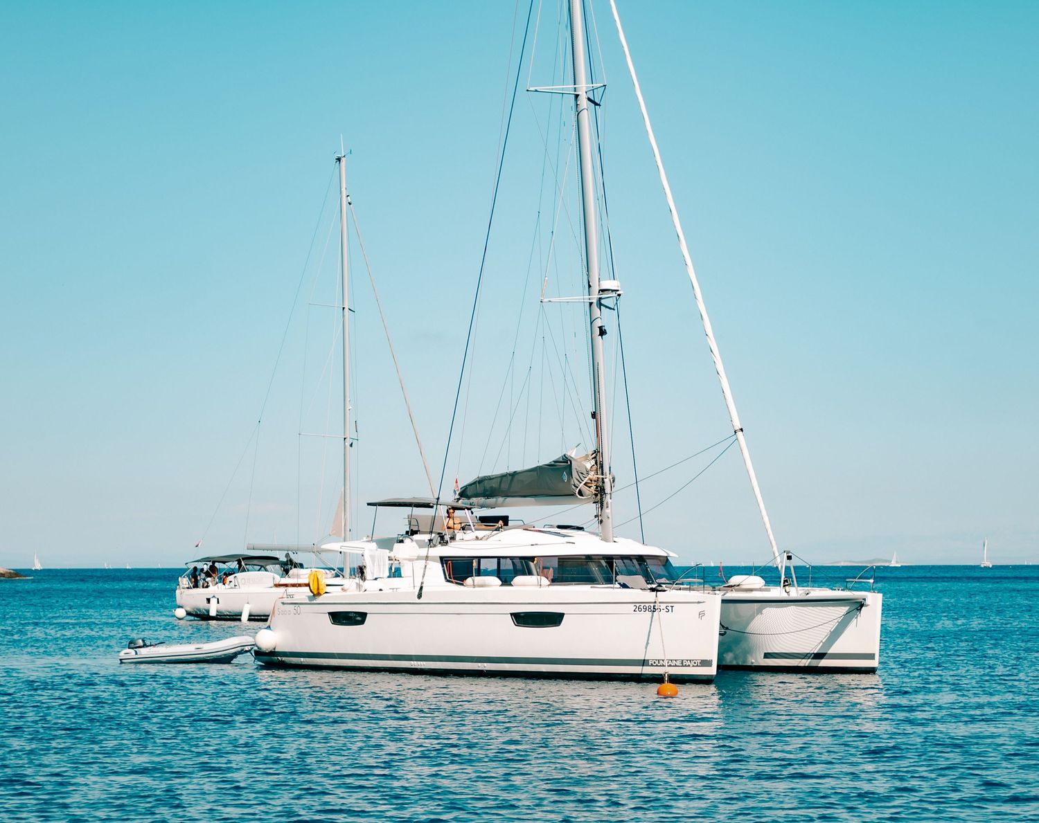 A white catamaran sailboat anchored on calm blue sea water under a clear sunny sky.