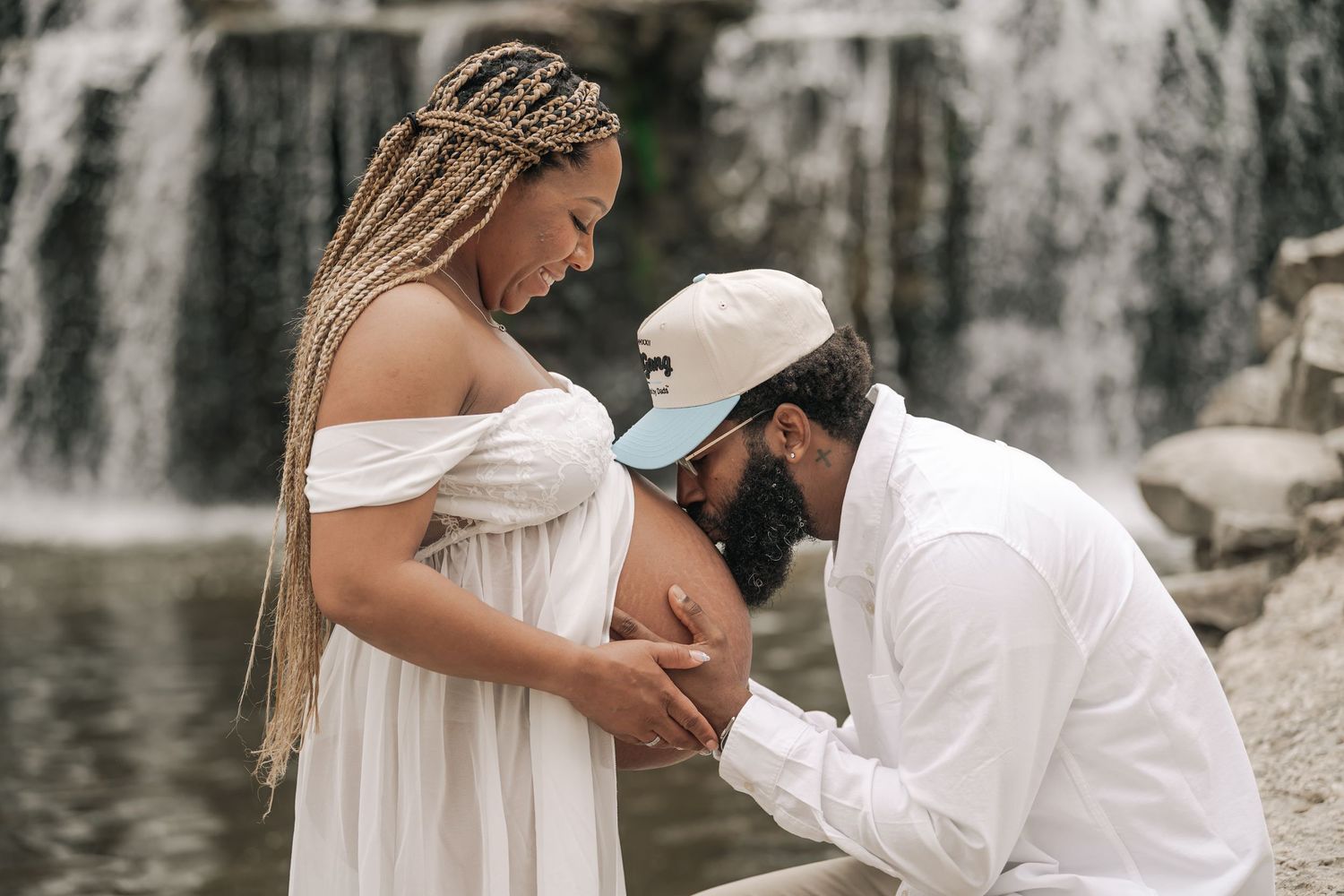 Man kneeling to kiss pregnant woman's belly during maternity shoot by a waterfall.