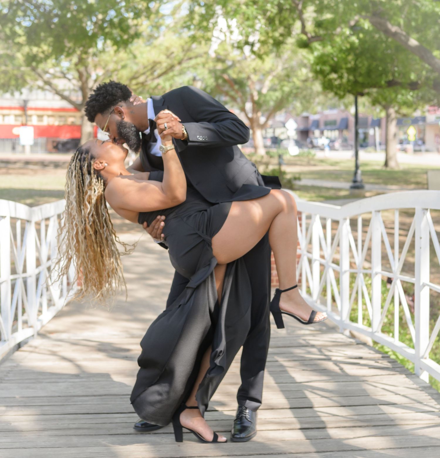 A stylish couple in black formal wear share a romantic dip on a white bridge outdoors.
