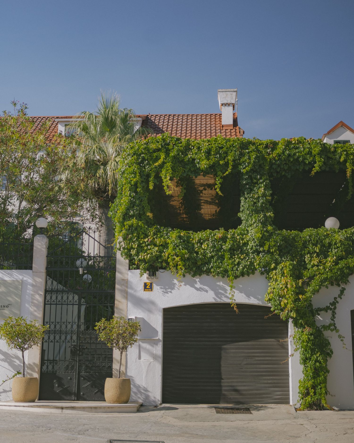 White Crotatian villa covered in lush green ivy with a garage and potted trees outside.