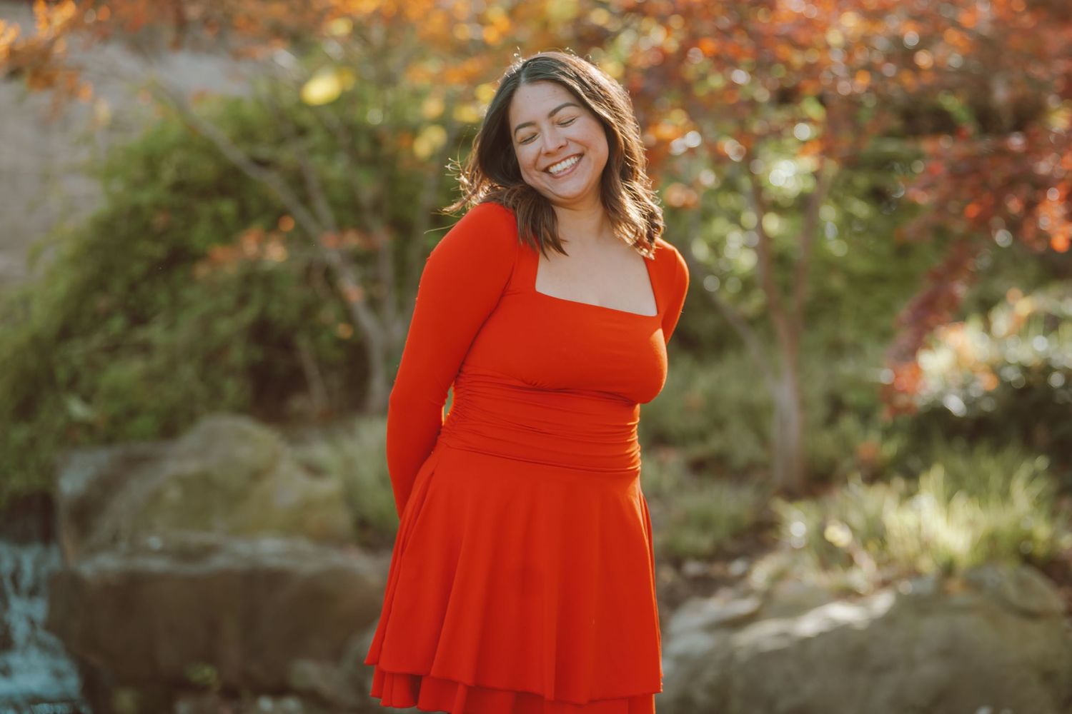 A smiling plus-size woman in a red dress poses outdoors among colorful autumn foliage.