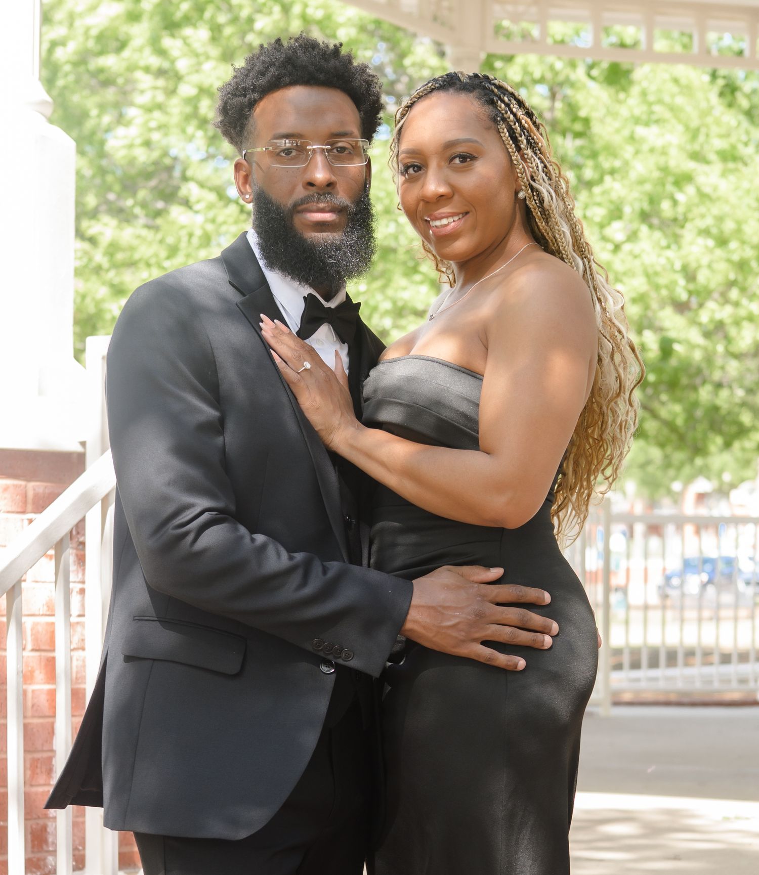 A well-dressed couple in black formal attire pose together under a shaded outdoor pavilion.