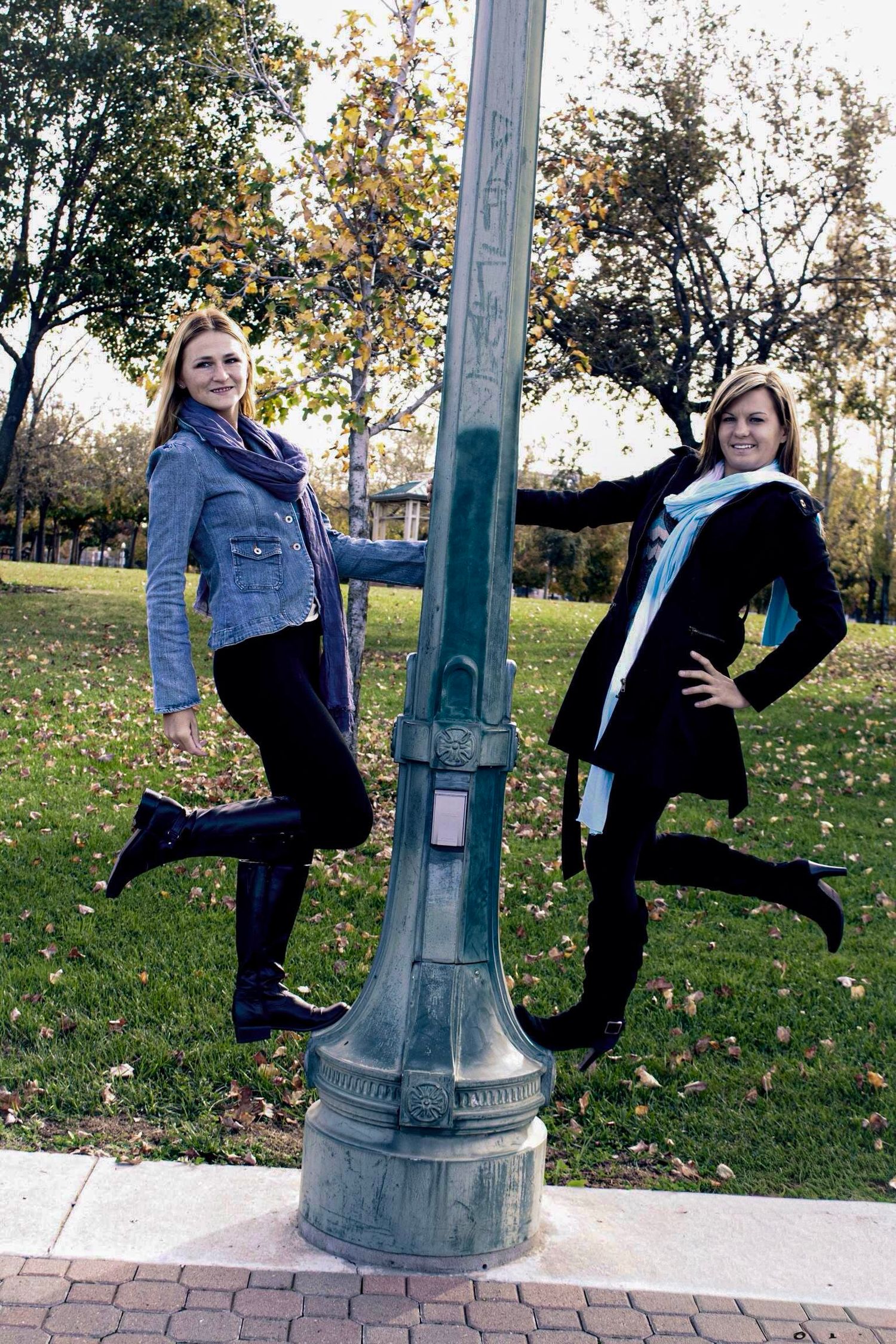 Two smiling young women jumping beside a green lamp post in an autumn park with colorful trees.