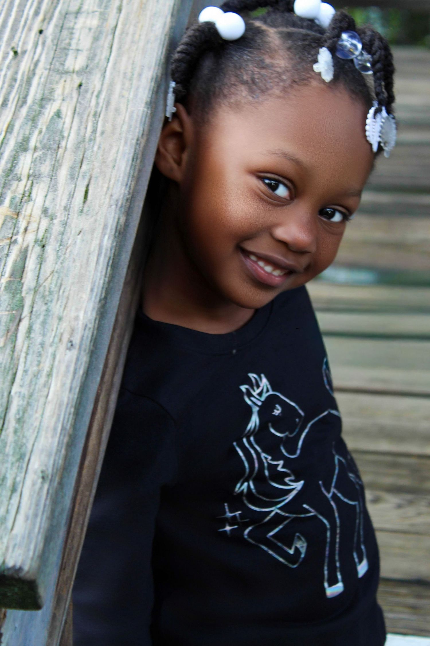 Smiling young Black girl with braids and white hair accessories wearing a black shirt, leaning against a wooden post.