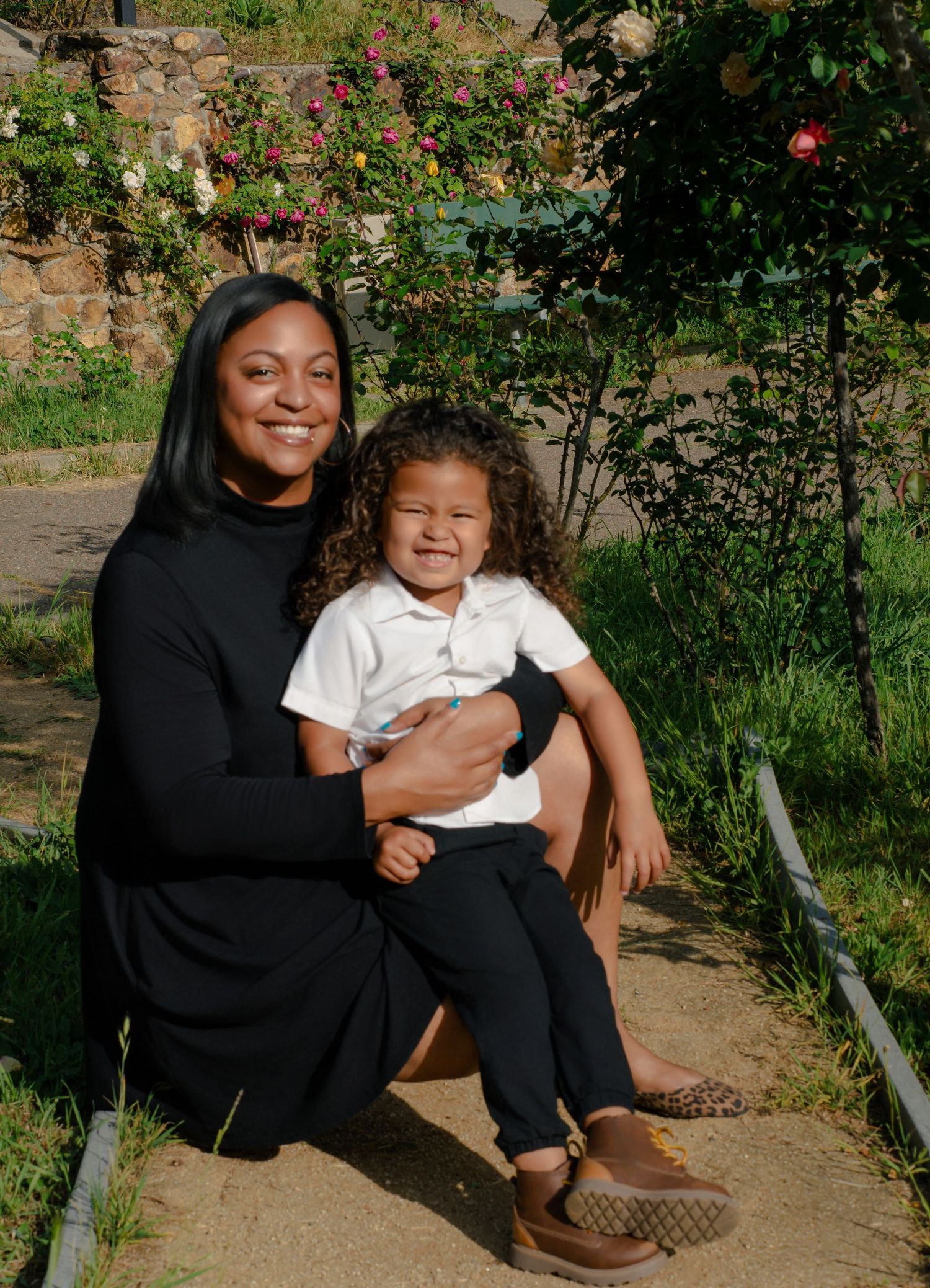 A smiling Black woman in a black dress sits outdoors holding a curly-haired child in a white polo shirt.